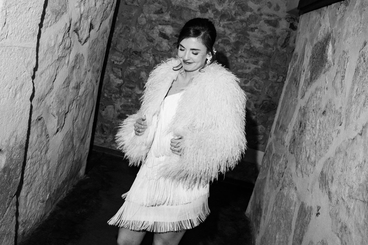 A figure in a white furry coat and fringed dress poses against stone walls in a black and white photograph.