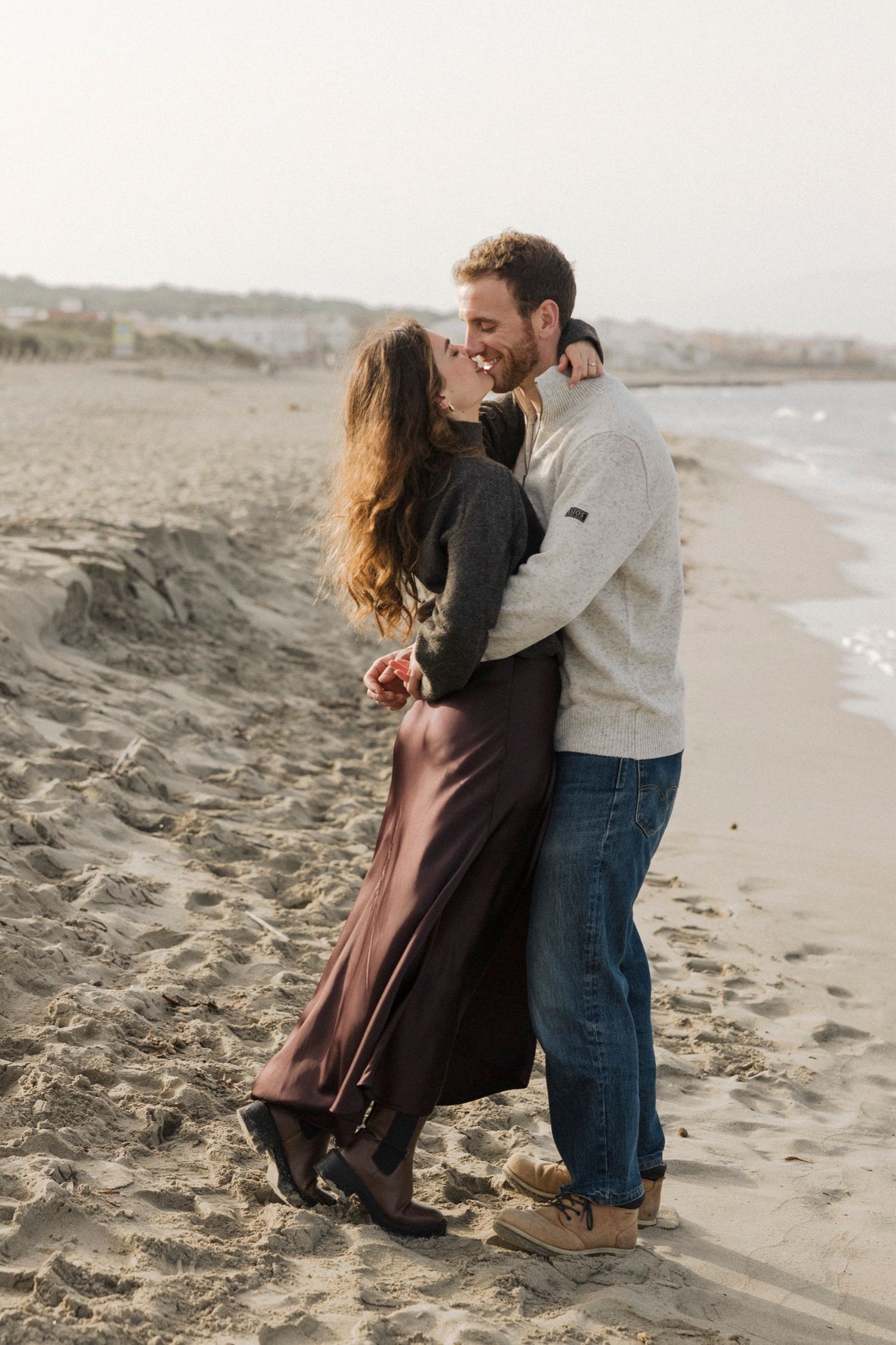 A couple shares a romantic moment together on a sandy beach at sunset with waves in the background.