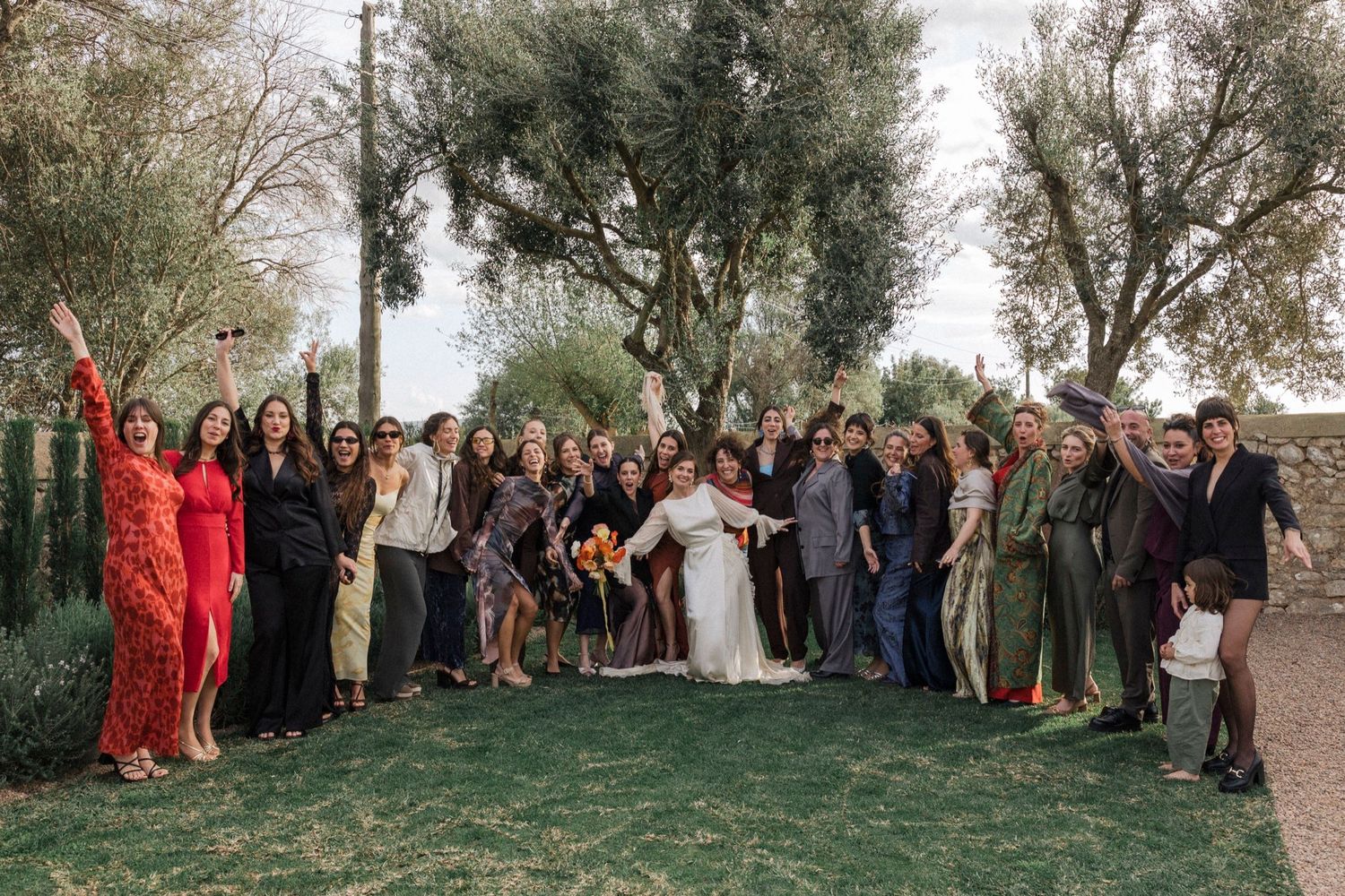 Large wedding party poses together outdoors in front of trees during an autumn celebration.