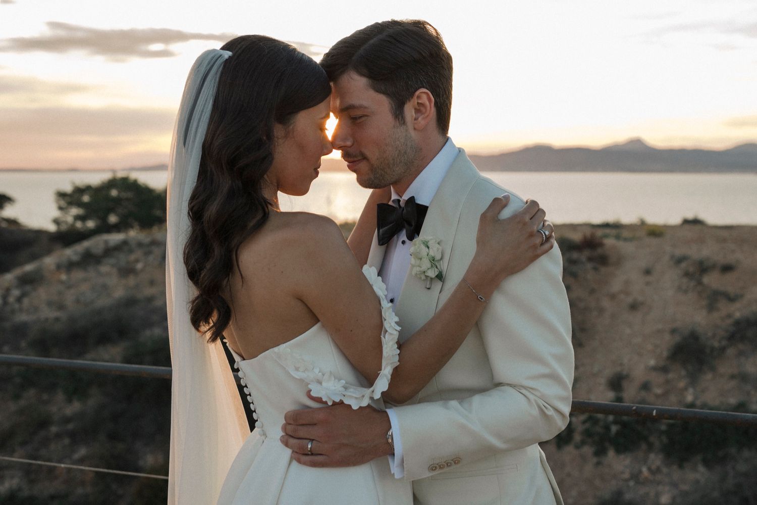 Bride and groom share romantic sunset moment outdoors, she in white gown with veil, he in ivory tuxedo with bow tie.