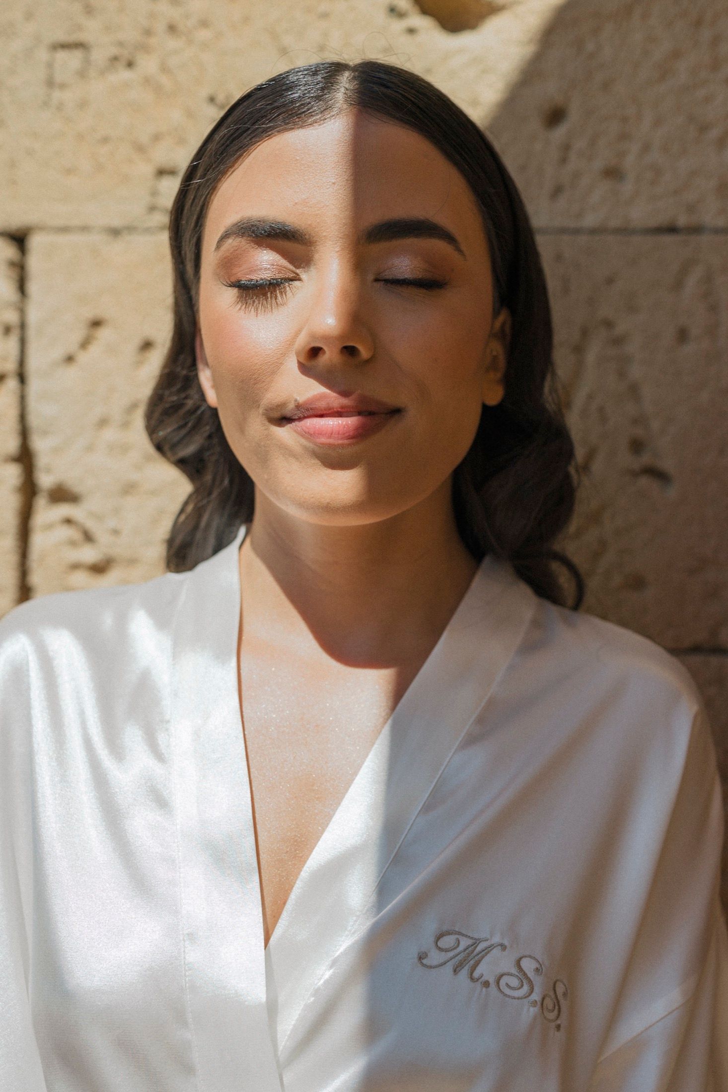 Bride in monogrammed white satin robe with eyes closed, wearing elegant makeup, posing against a stone wall.
