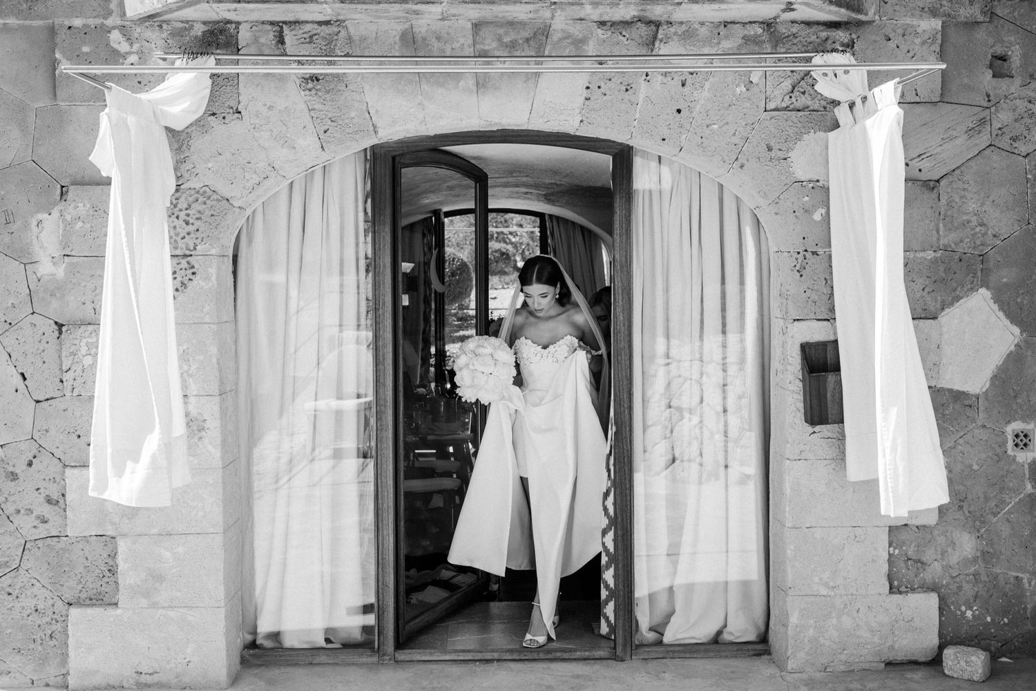 Bride in white dress standing in stone doorway with flowing curtains, elegant black and white wedding photo.