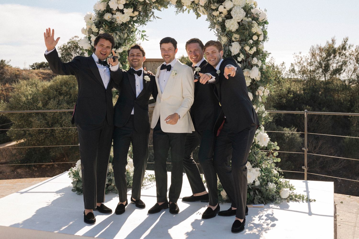 Groom in white tuxedo jacket poses with four groomsmen in black suits at an outdoor wedding ceremony with floral arch.