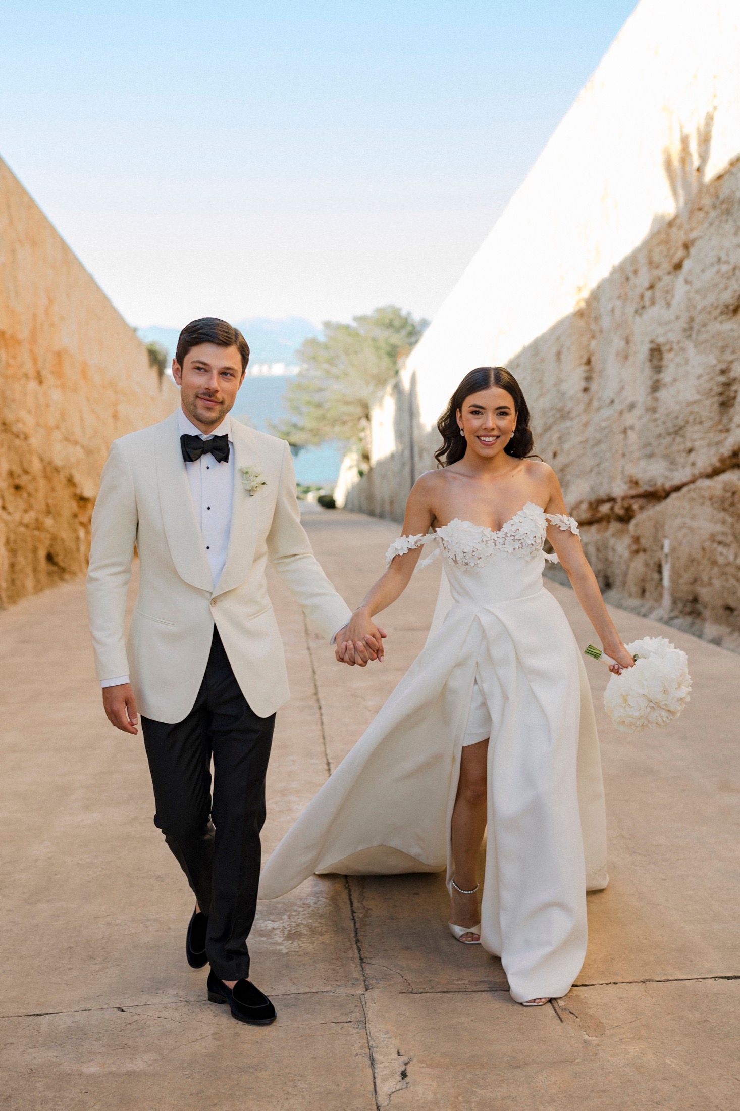 A smiling bride and groom walk hand-in-hand outdoors, she in a white off-shoulder gown, he in a cream tuxedo.
