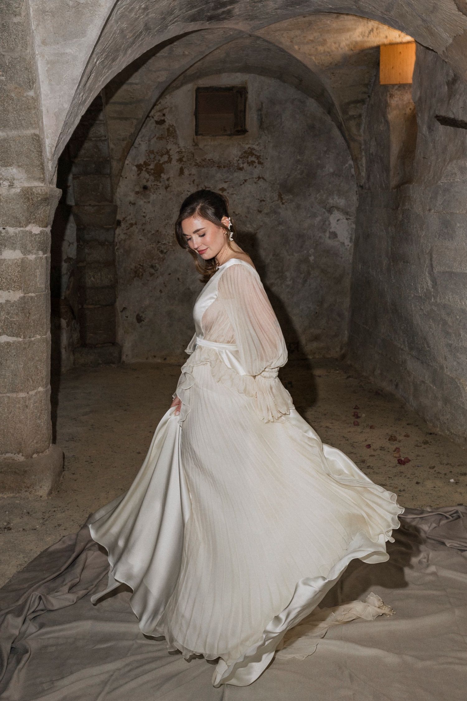 A person in a flowing white vintage-style dress poses dramatically in an atmospheric stone basement or crypt setting.