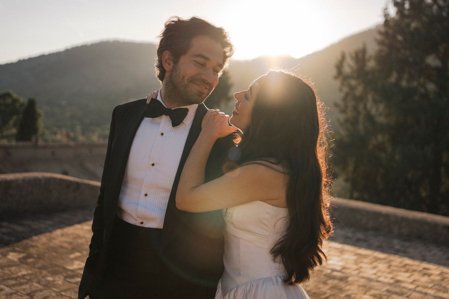 A romantic couple embraces during golden hour in an outdoor setting with mountains and sunlight in the background.