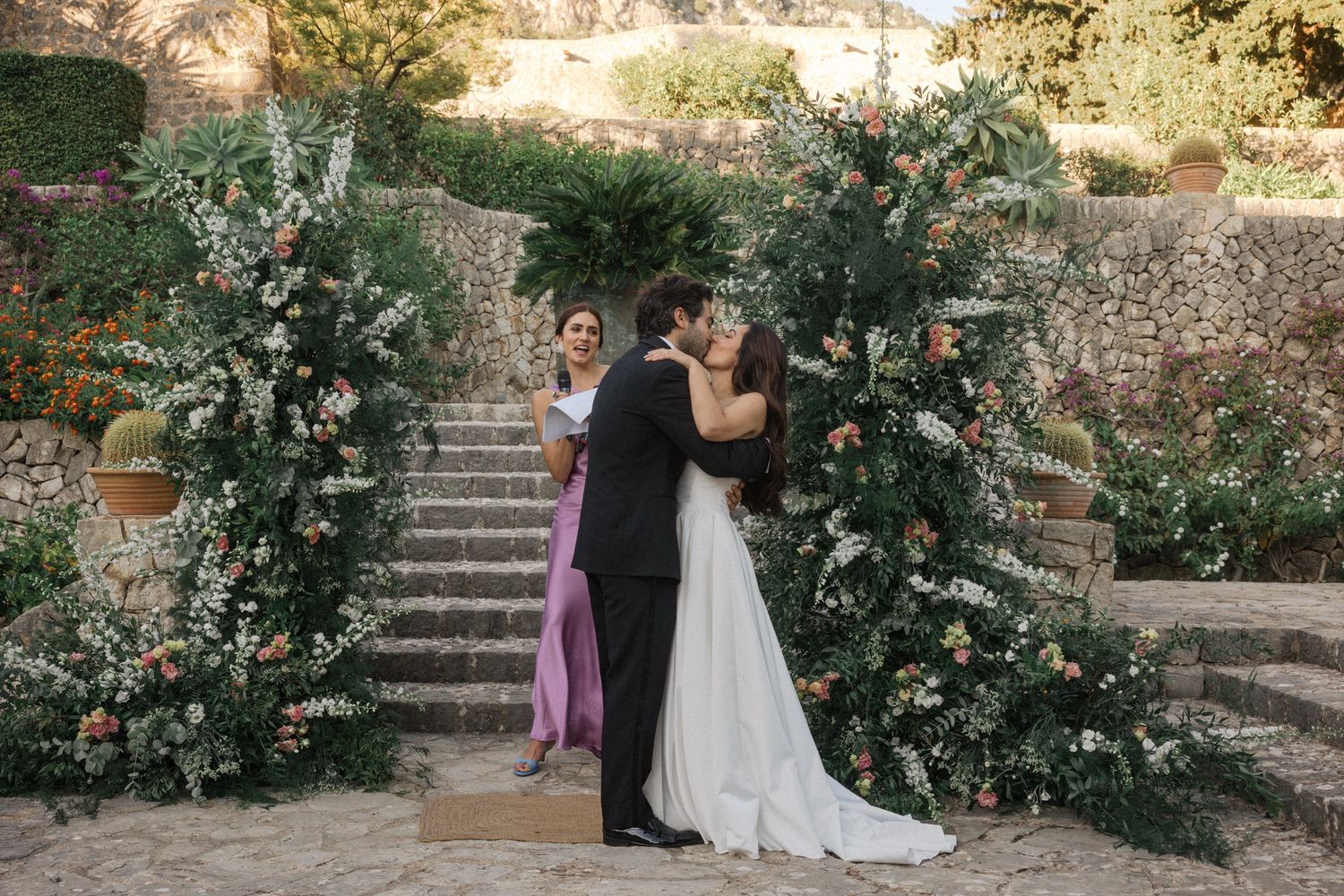 A romantic wedding ceremony takes place in a lush garden setting with stone steps and natural landscaping.
