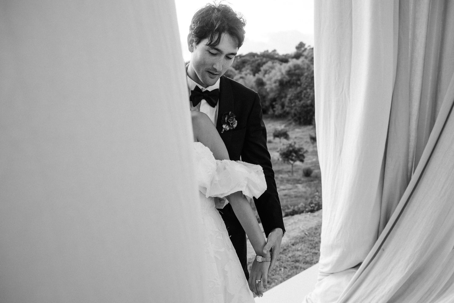 A black and white wedding photo showing a tender moment between a couple behind white curtains outdoors.