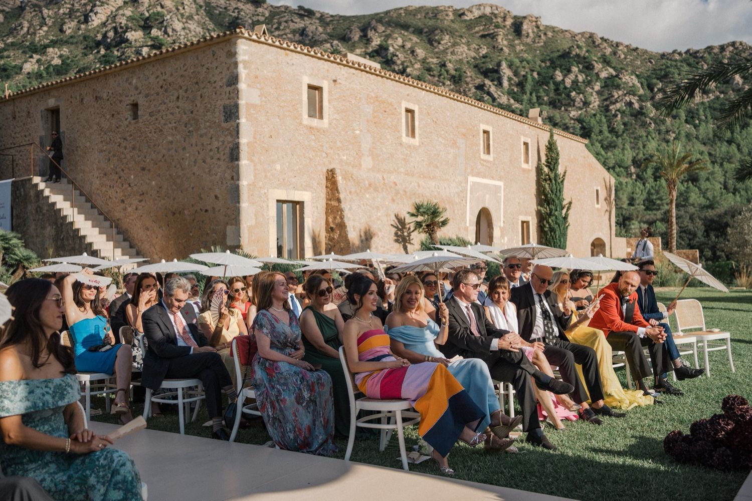Guests seated in white chairs at an outdoor event in front of a rustic stone building in a mountainous setting.