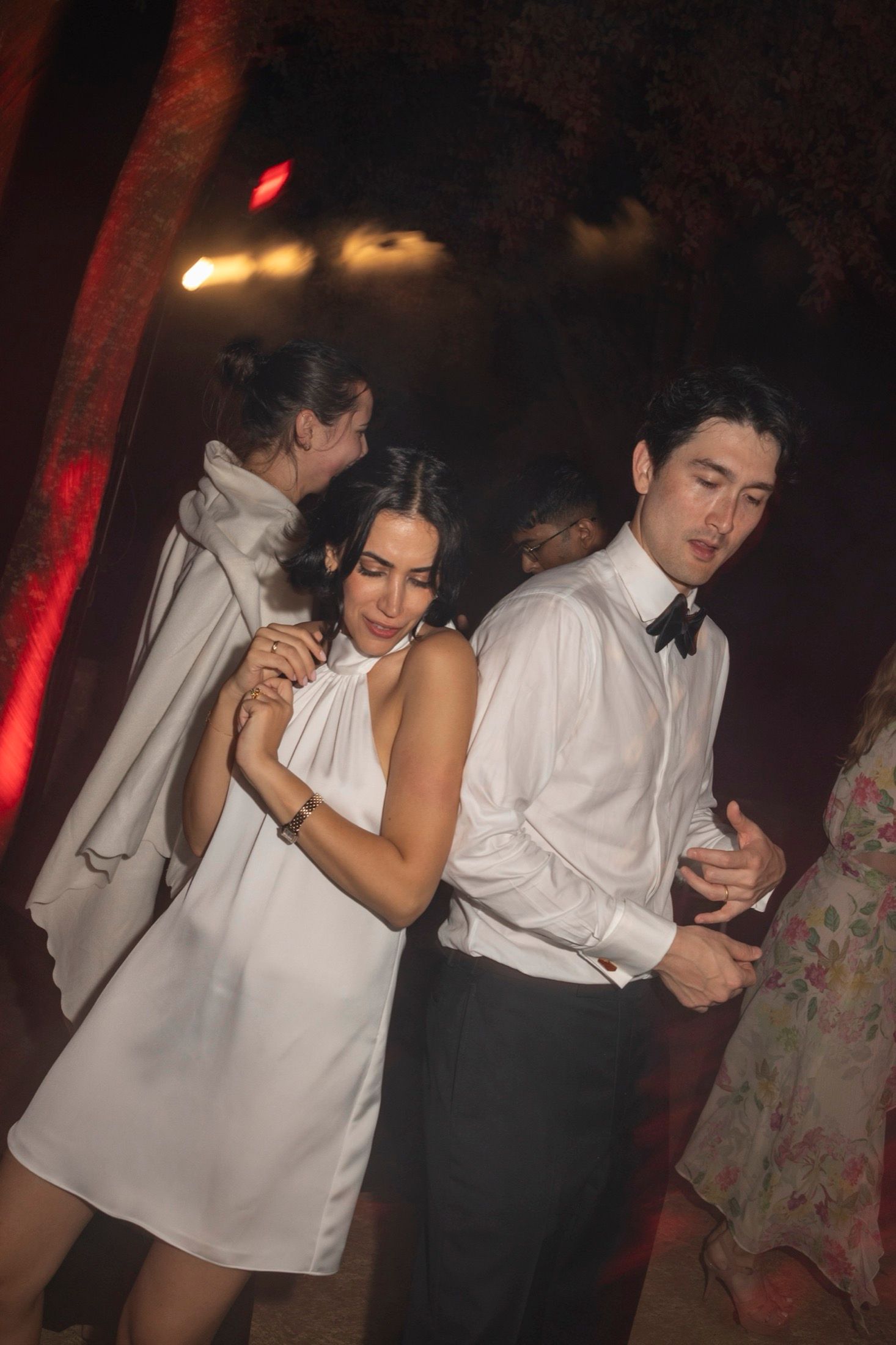 People in formal attire dance at a dimly lit party with red lighting and atmospheric ambiance.