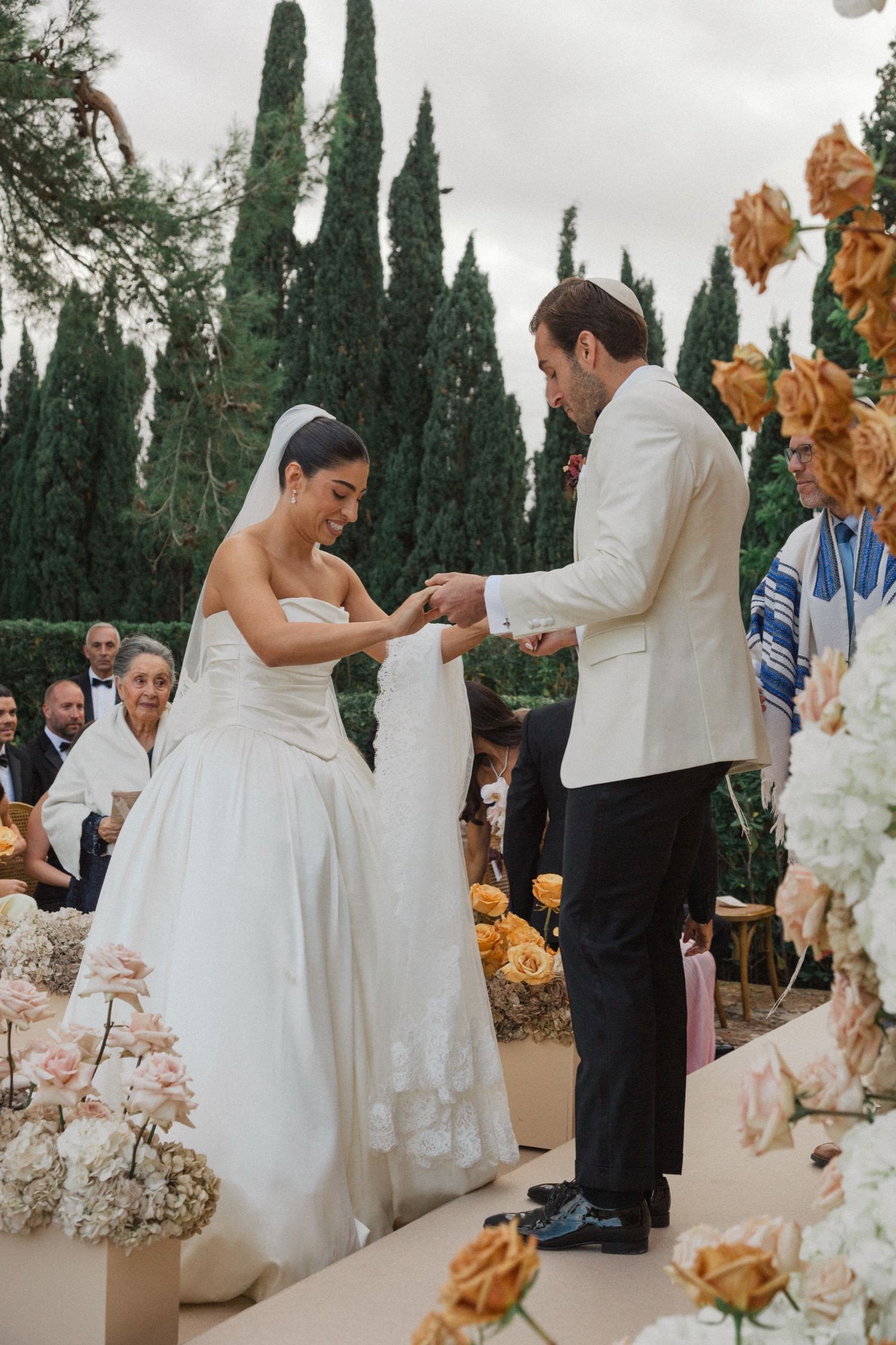 A wedding ceremony takes place outdoors with tall cypress trees in the background and floral decorations along the aisle.