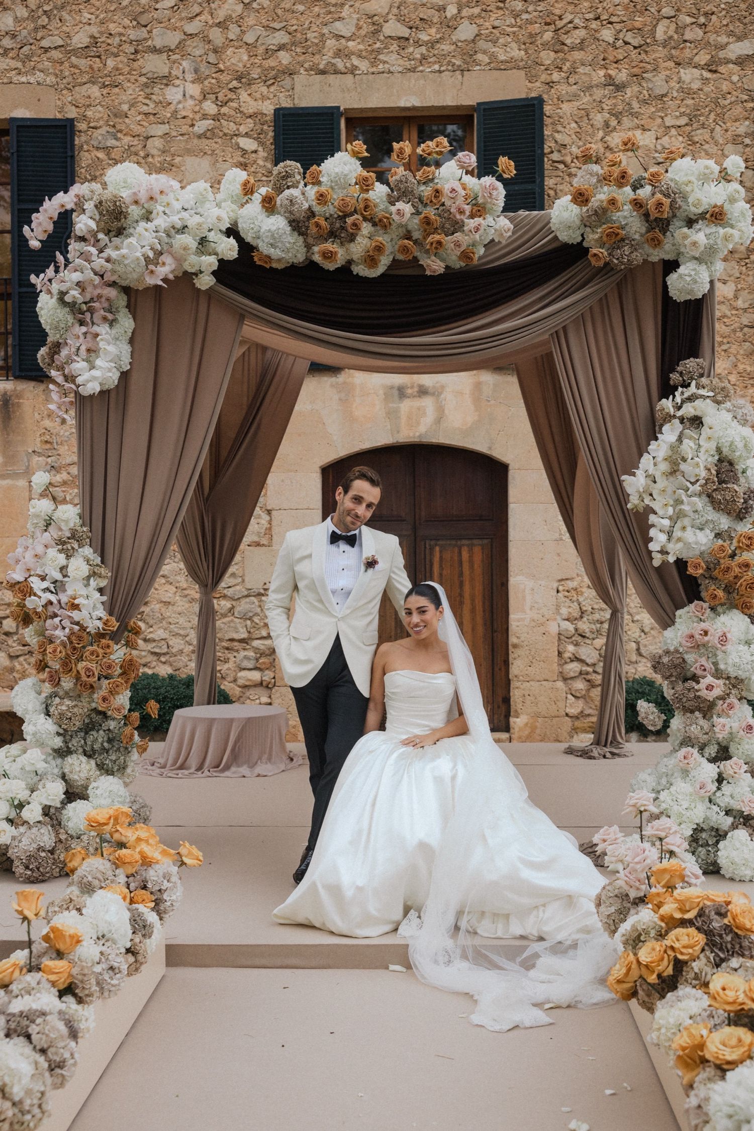 Elegant wedding scene with brown drapes and floral arches featuring white and peach roses at a rustic stone venue.