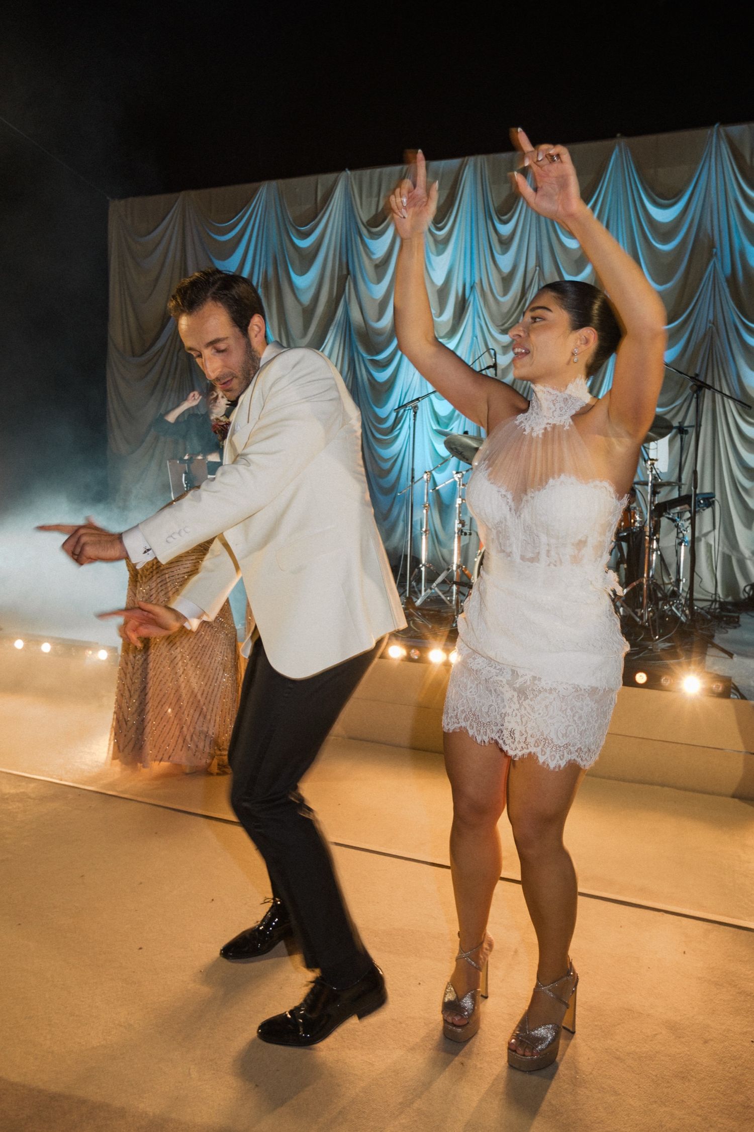 Bride in short lace dress and groom in white jacket dance energetically on wedding dance floor with blue backdrop.