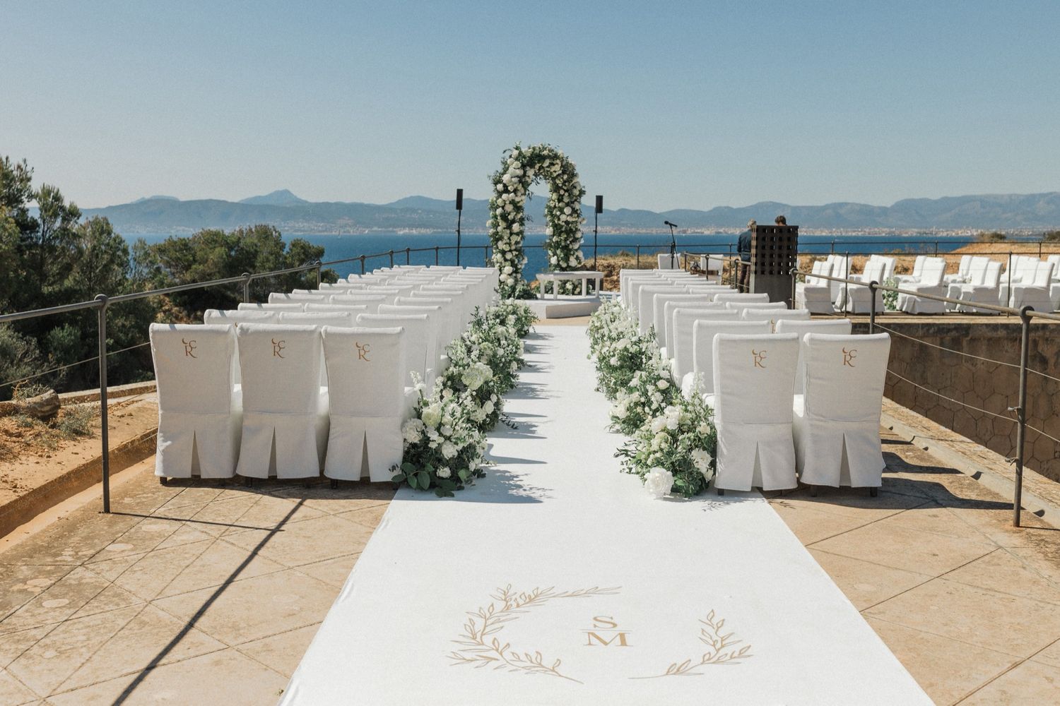 Elegant outdoor wedding ceremony setup with white chairs, floral arch, and aisle runner overlooking a scenic ocean view.
