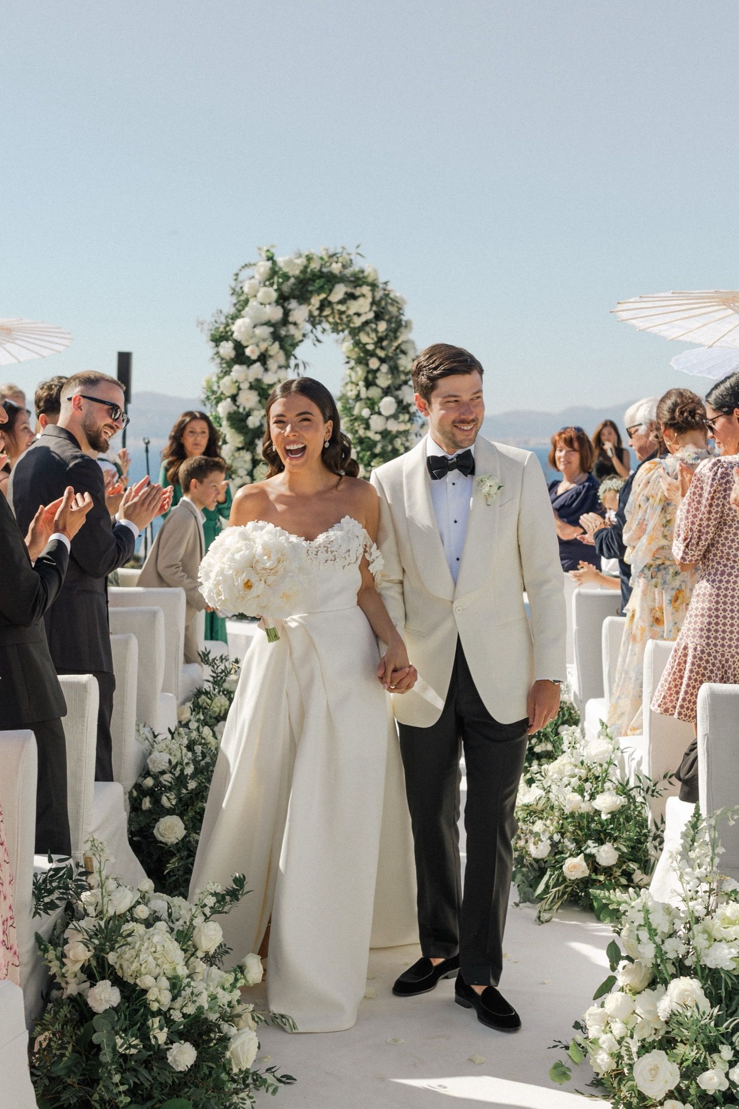 Bride and groom walk down the aisle at an outdoor wedding ceremony with white floral arch.