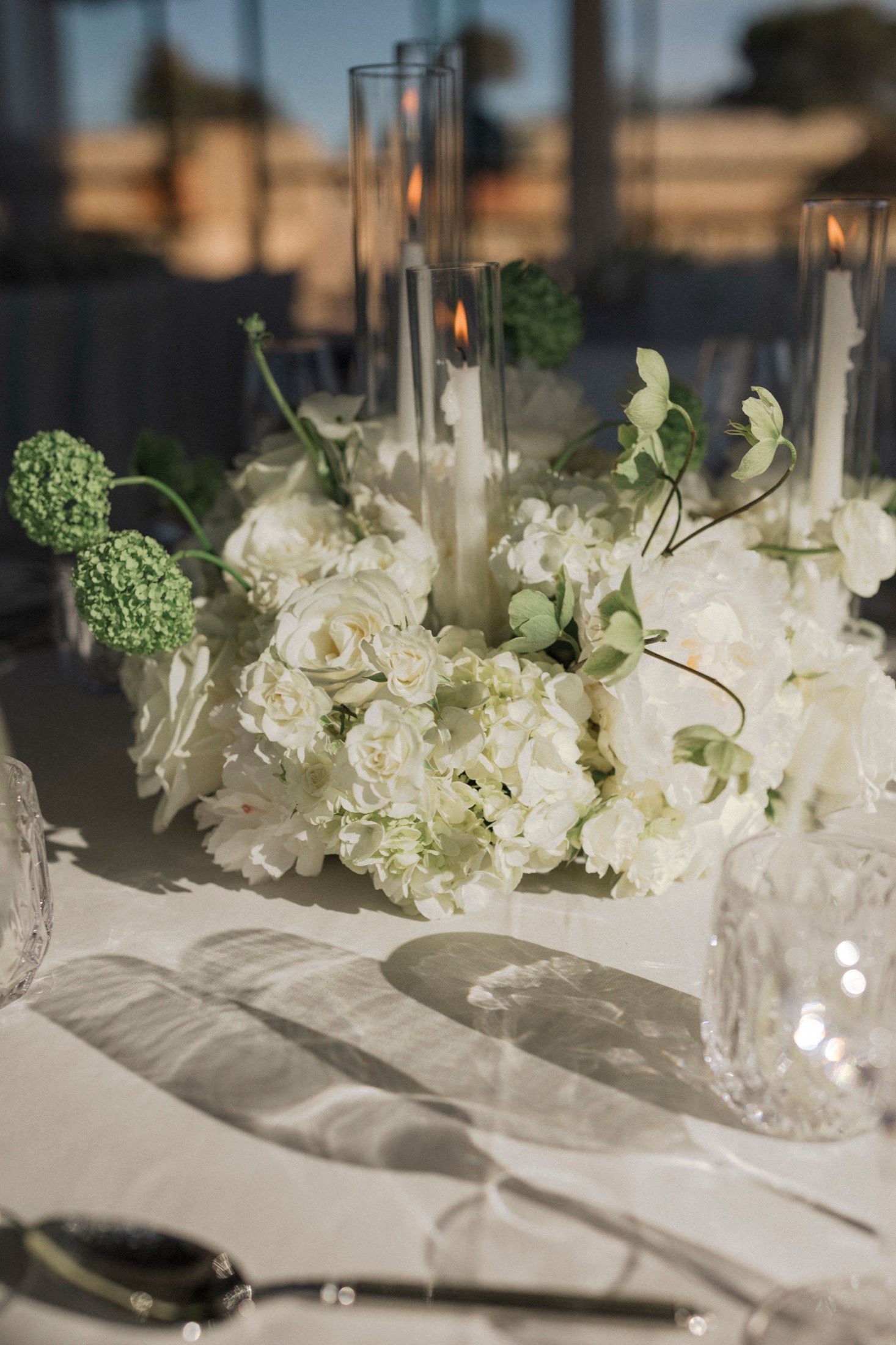 Elegant wedding table centerpiece with white hydrangeas, roses, and lit candles in glass holders on a white linen tablecloth.