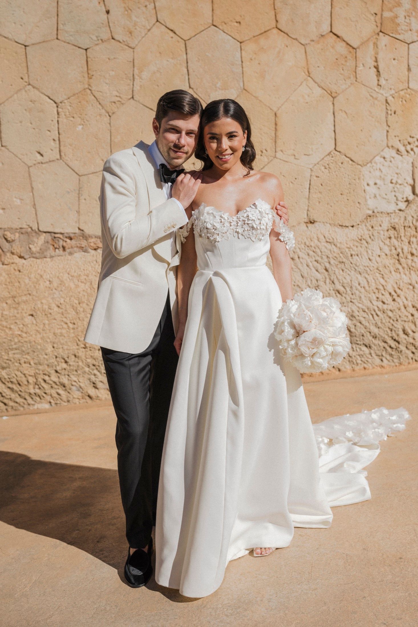 Bride in floral off-shoulder gown holds white bouquet beside groom in ivory tuxedo against stone wall.