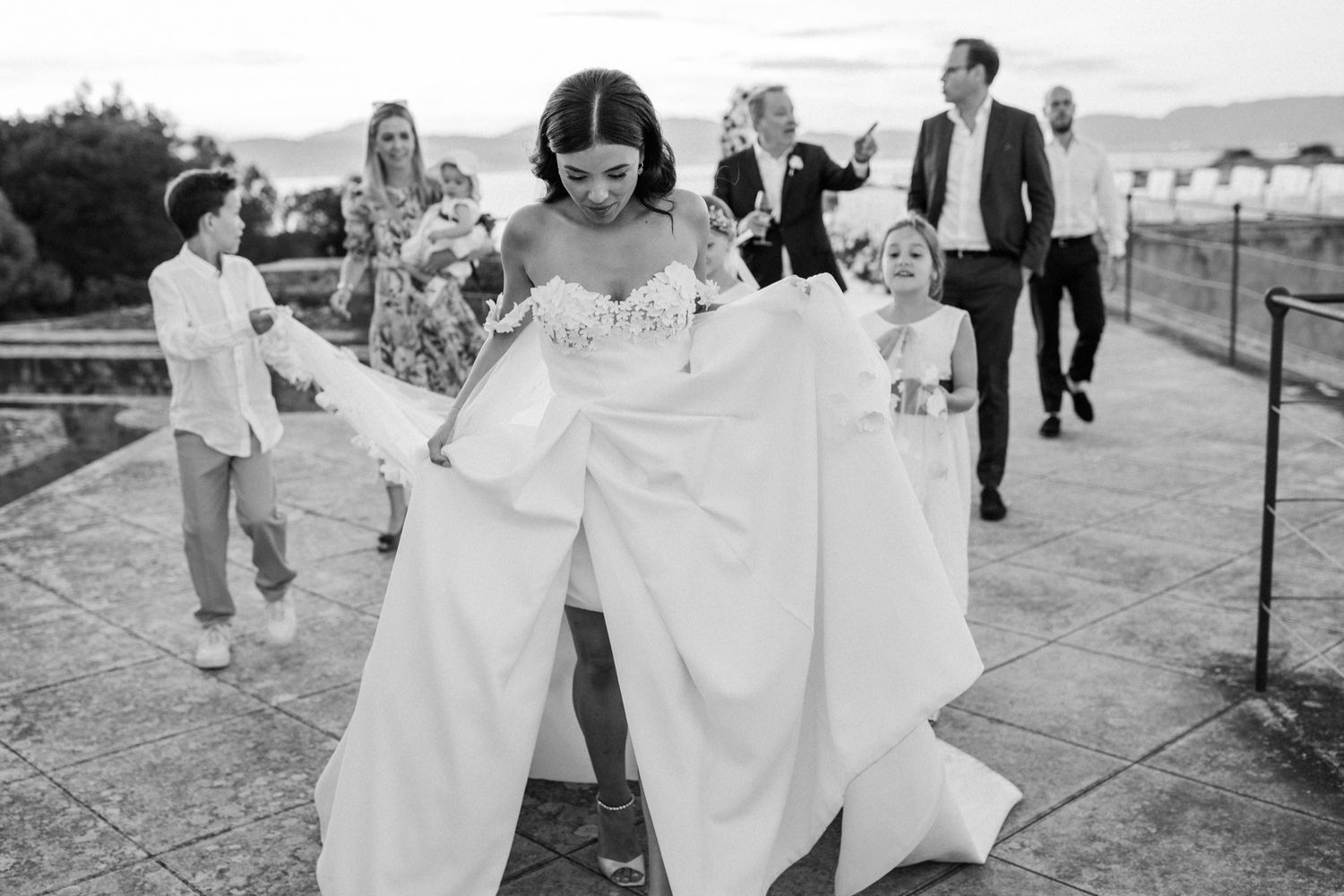 Bride in white gown lifts dress while walking outdoors with wedding guests in black and white photo.
