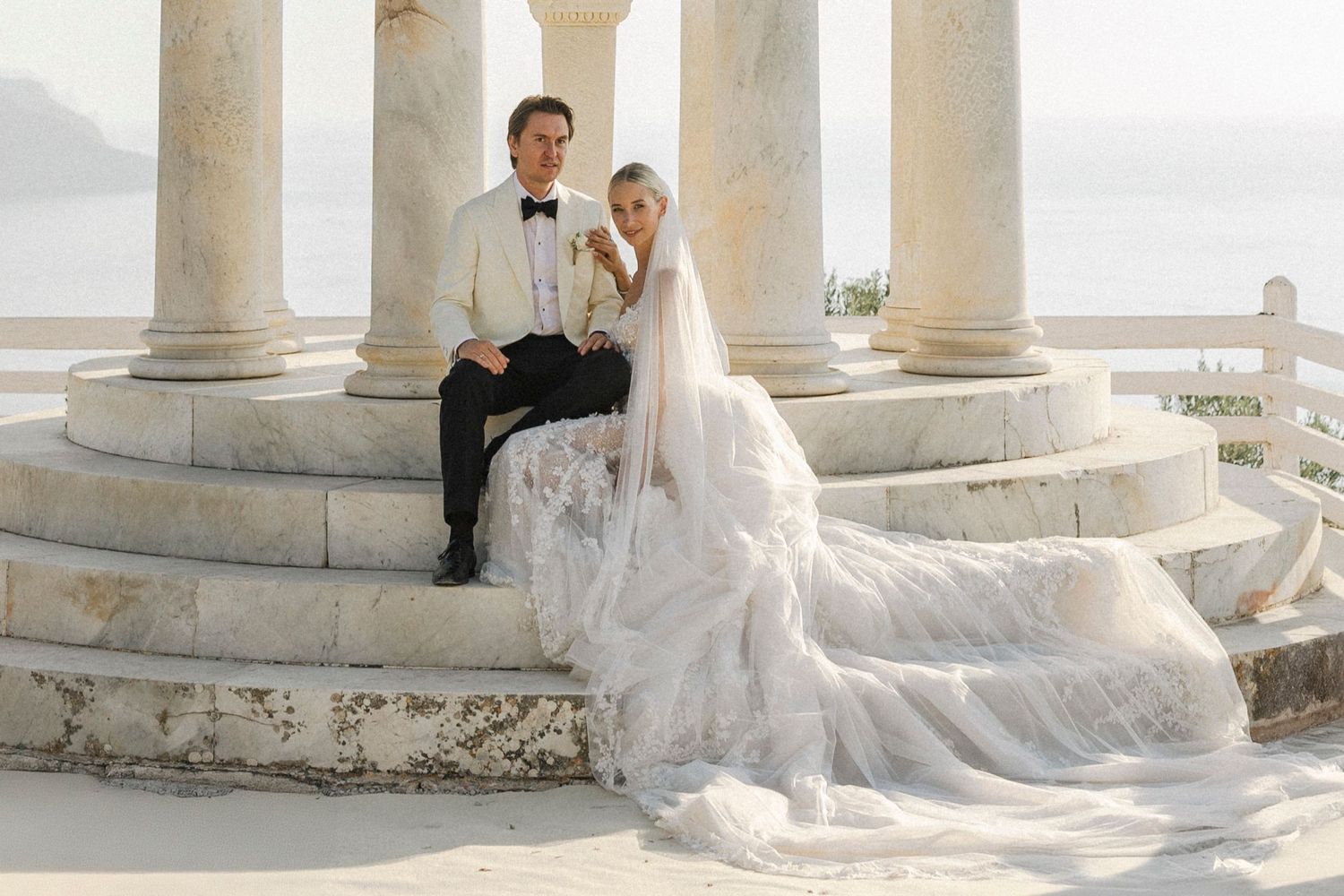 A bride in a flowing white wedding gown poses elegantly between classic stone columns at a seaside location.