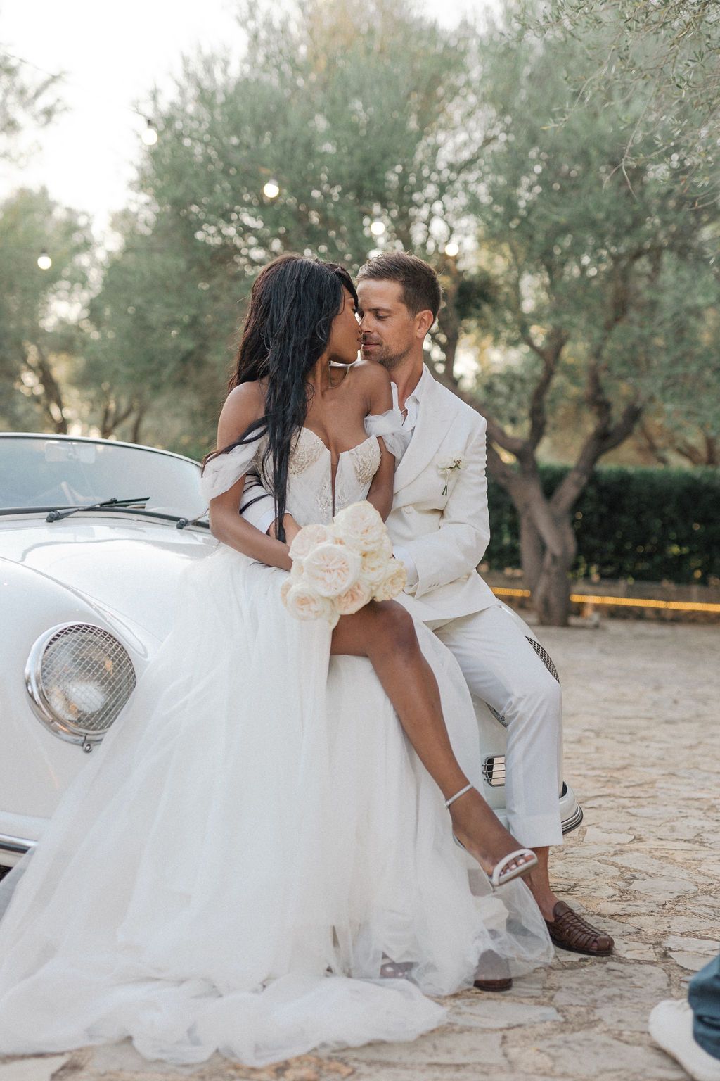 Newlyweds pose together on stone ground near vintage white automobile.