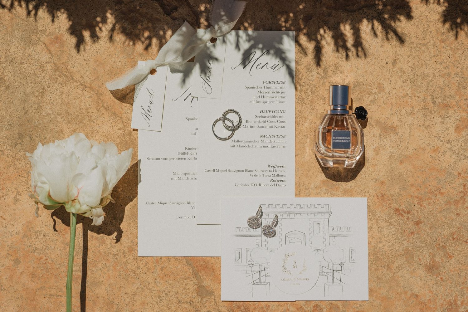 Wedding flat lay featuring invitation suite, gold rings, white peony, blue perfume bottle, and elegant stationery on warm stone.