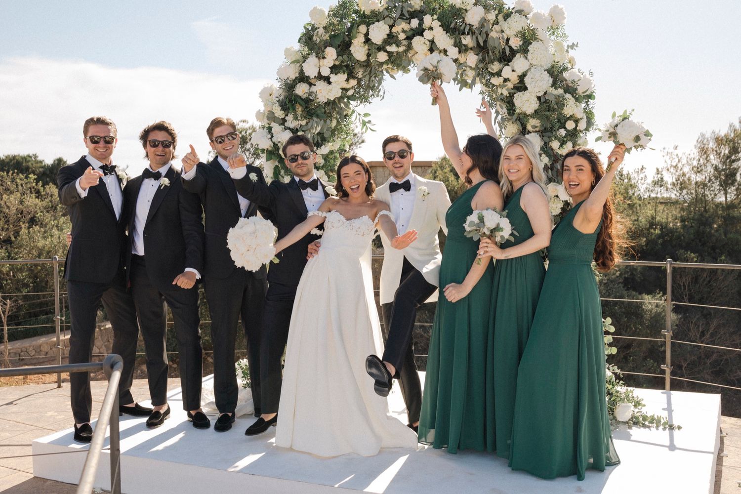 Wedding party celebrating outdoors with bride and groom, groomsmen in black suits and bridesmaids in green dresses near floral arch.
