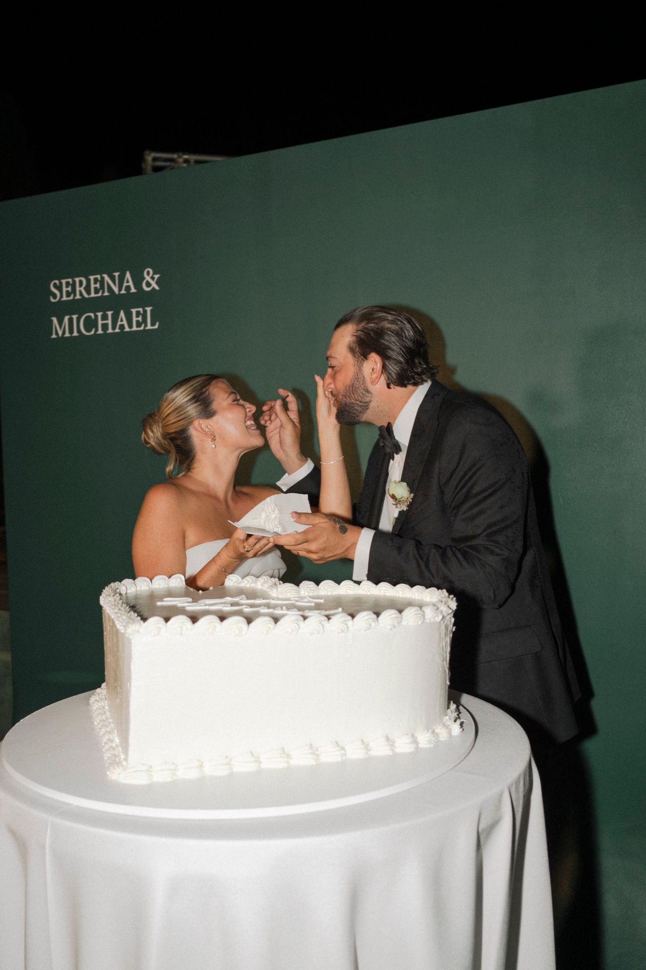 Couple leans over table to share cake at their wedding reception.