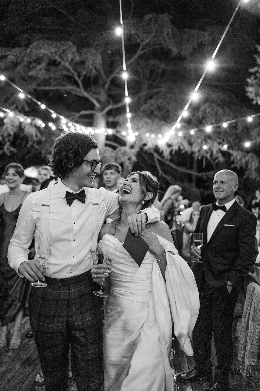 Wedding party dancing and laughing together under string lights at outdoor reception.