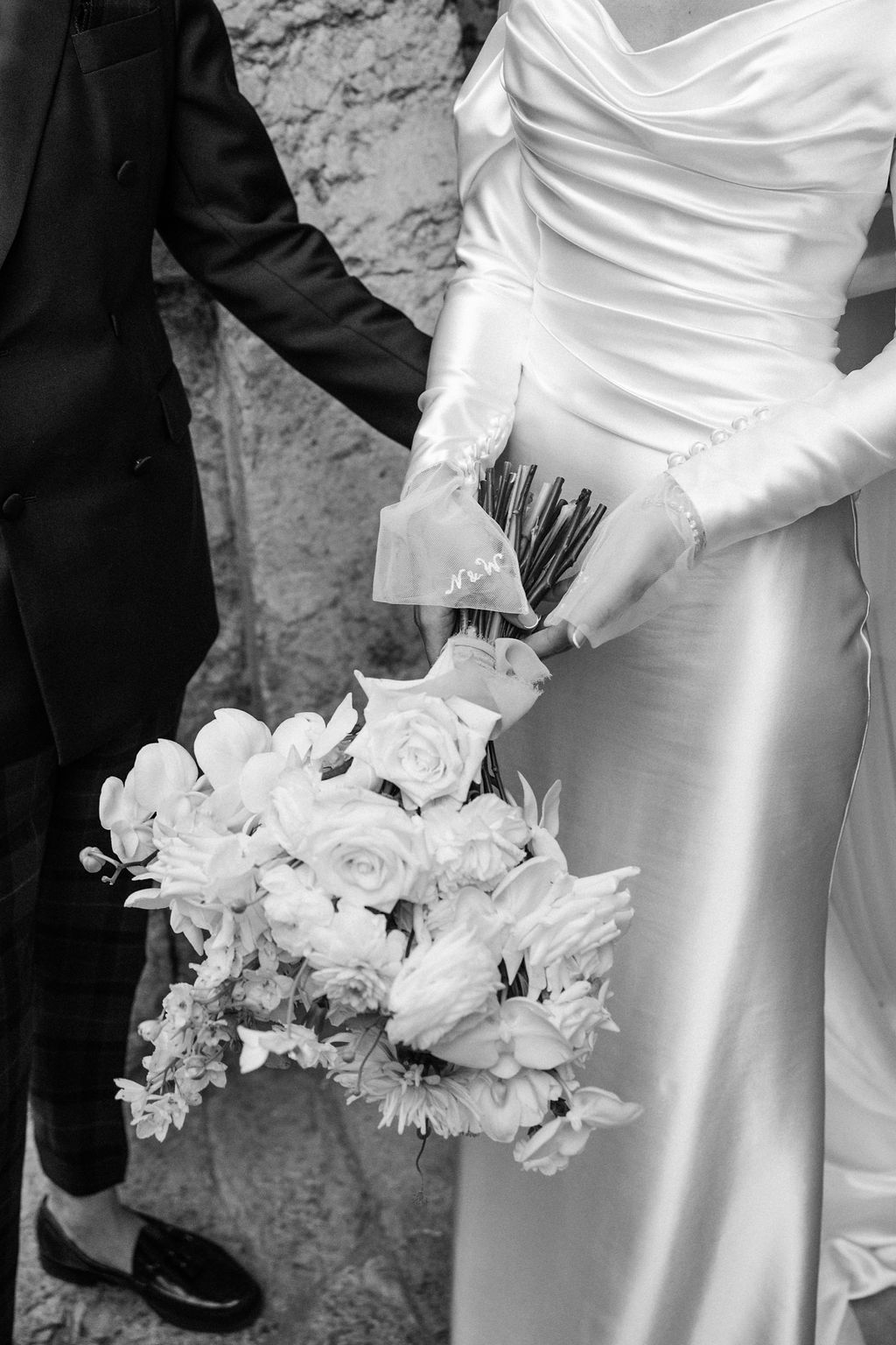 Close-up details of white wedding bouquet and elegant bridal gown against stone wall backdrop.