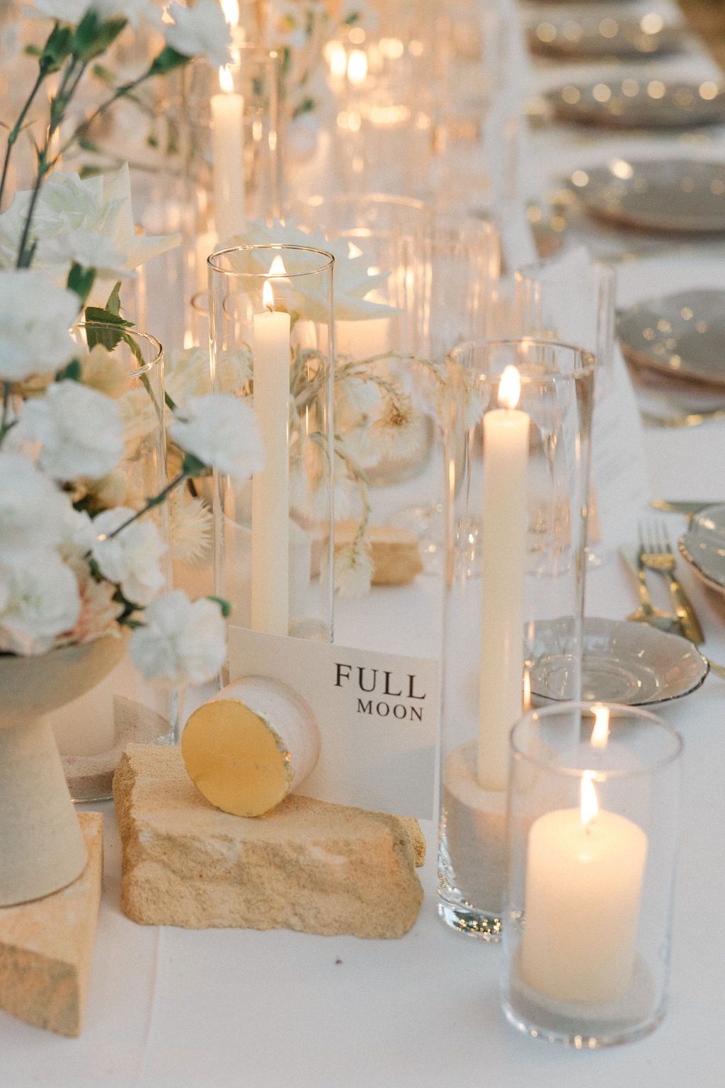 Elegant wedding table decor with glowing candles in glass cylinders and white floral arrangements.