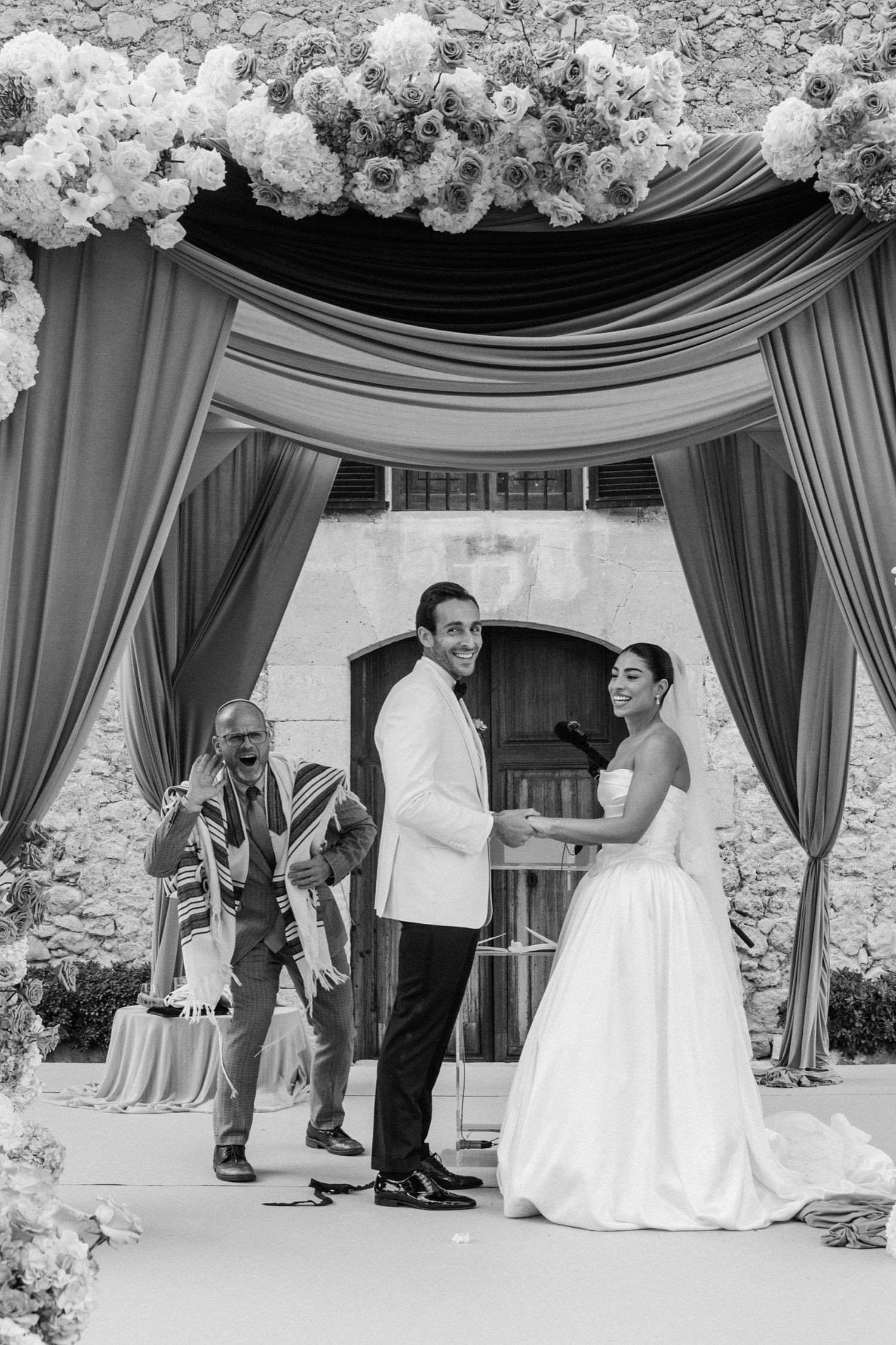 A black and white wedding ceremony photo showing a couple under an elegant draped canopy adorned with white flowers.