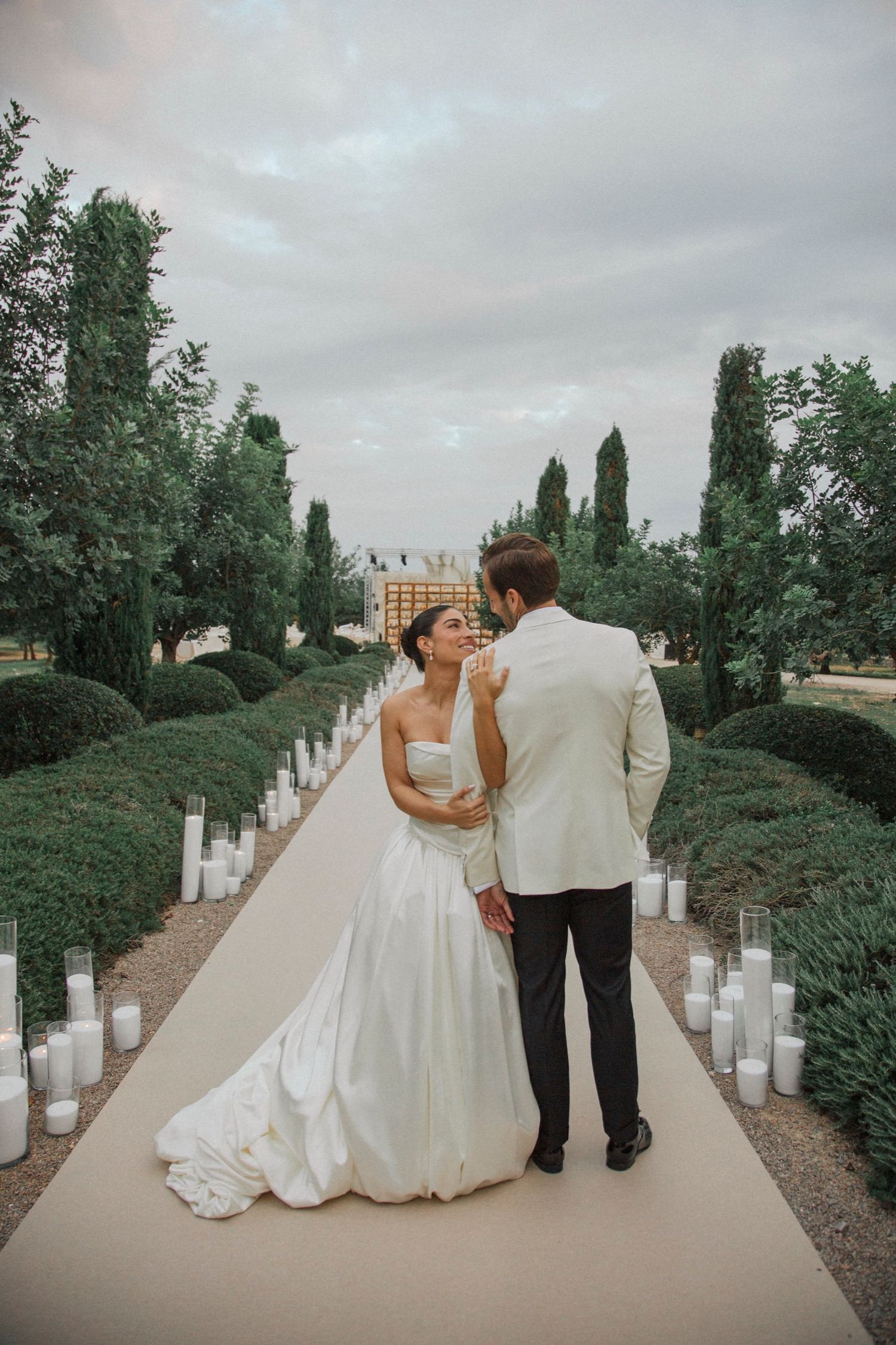 A romantic wedding couple walks along a garden path lined with glowing candles and tall cypress trees at dusk.