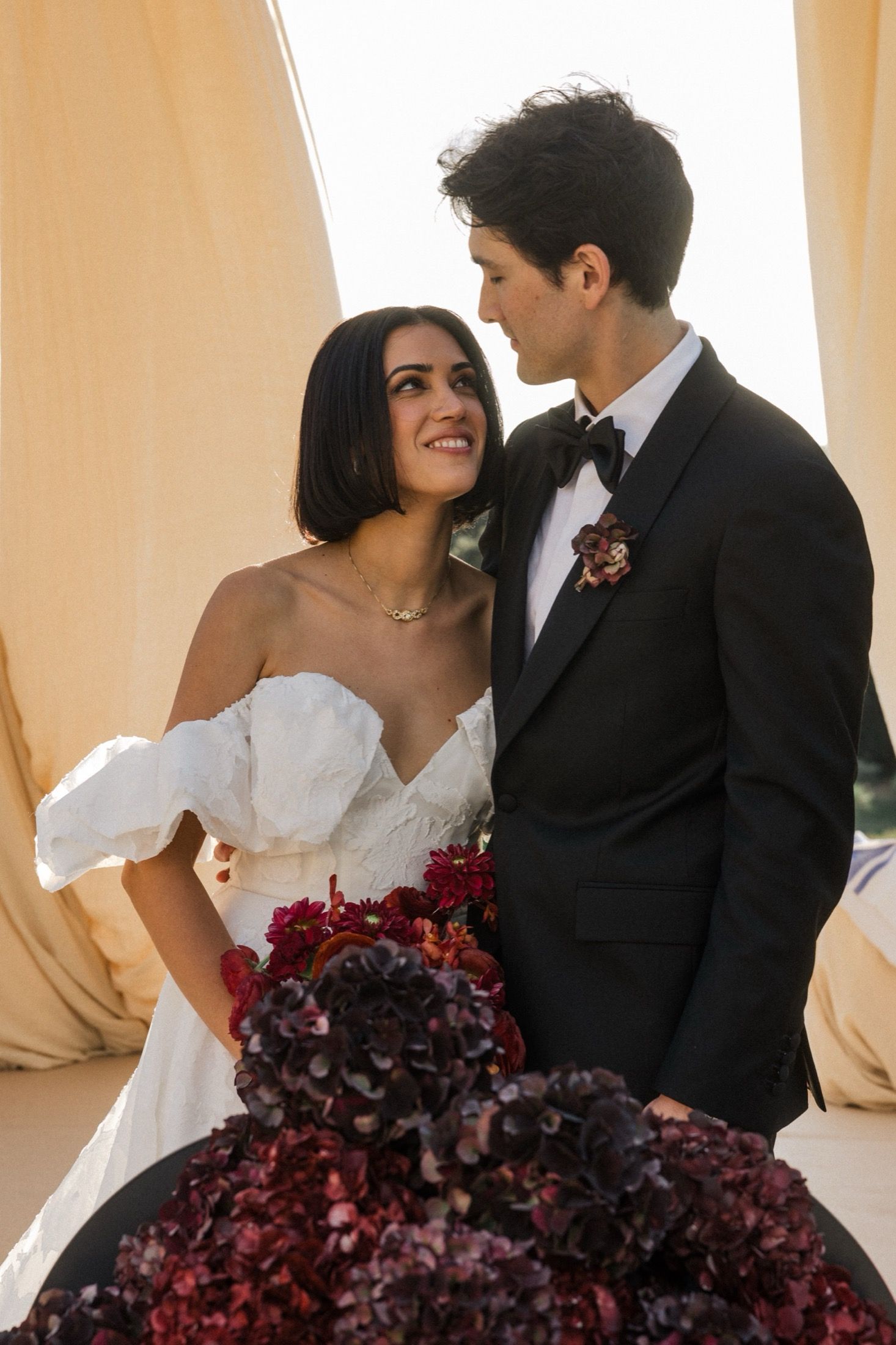 A bride in a white off-shoulder gown and groom in black tuxedo share an intimate moment with deep red floral bouquet.