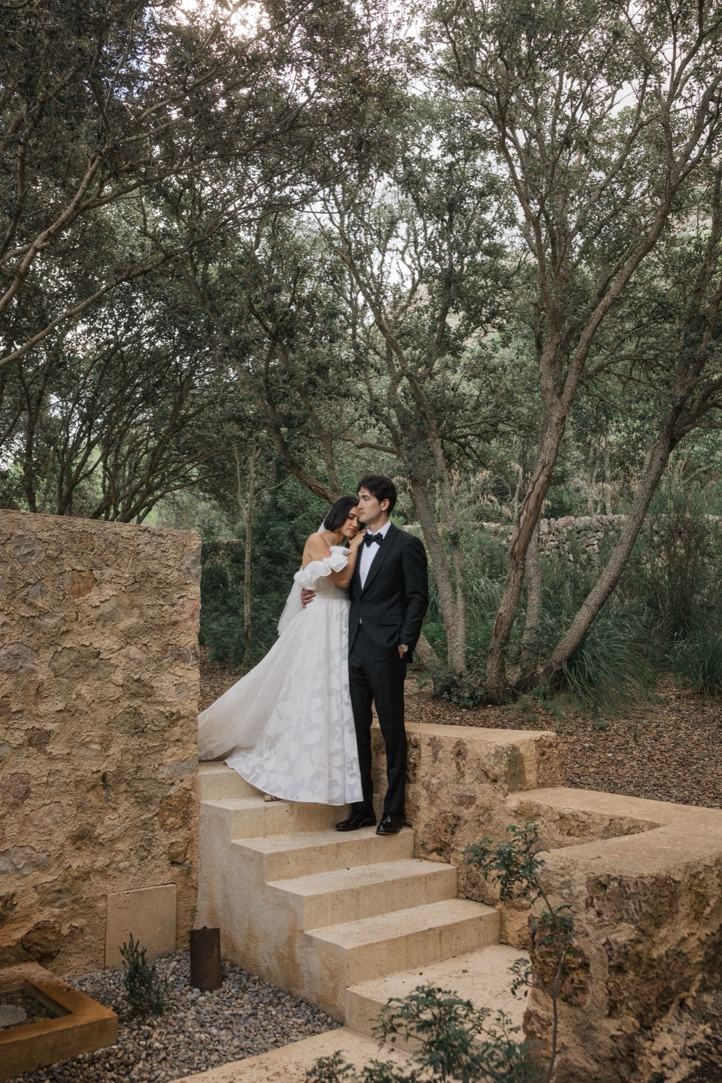 A couple in wedding attire share a romantic moment on stone steps surrounded by a wooded natural setting.