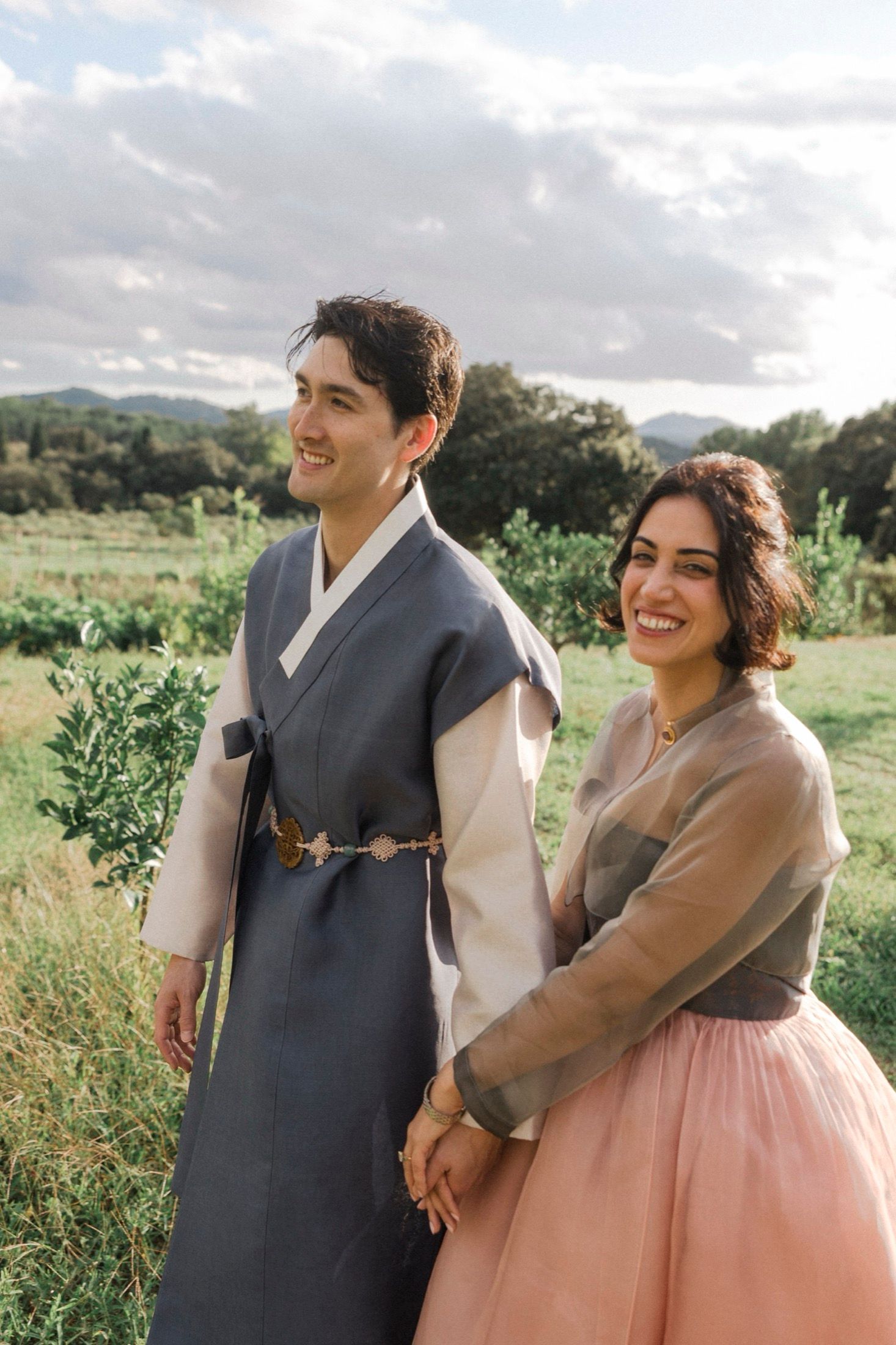 A couple in traditional Korean hanbok dress laughing together in a sunny outdoor field with mountains in background.