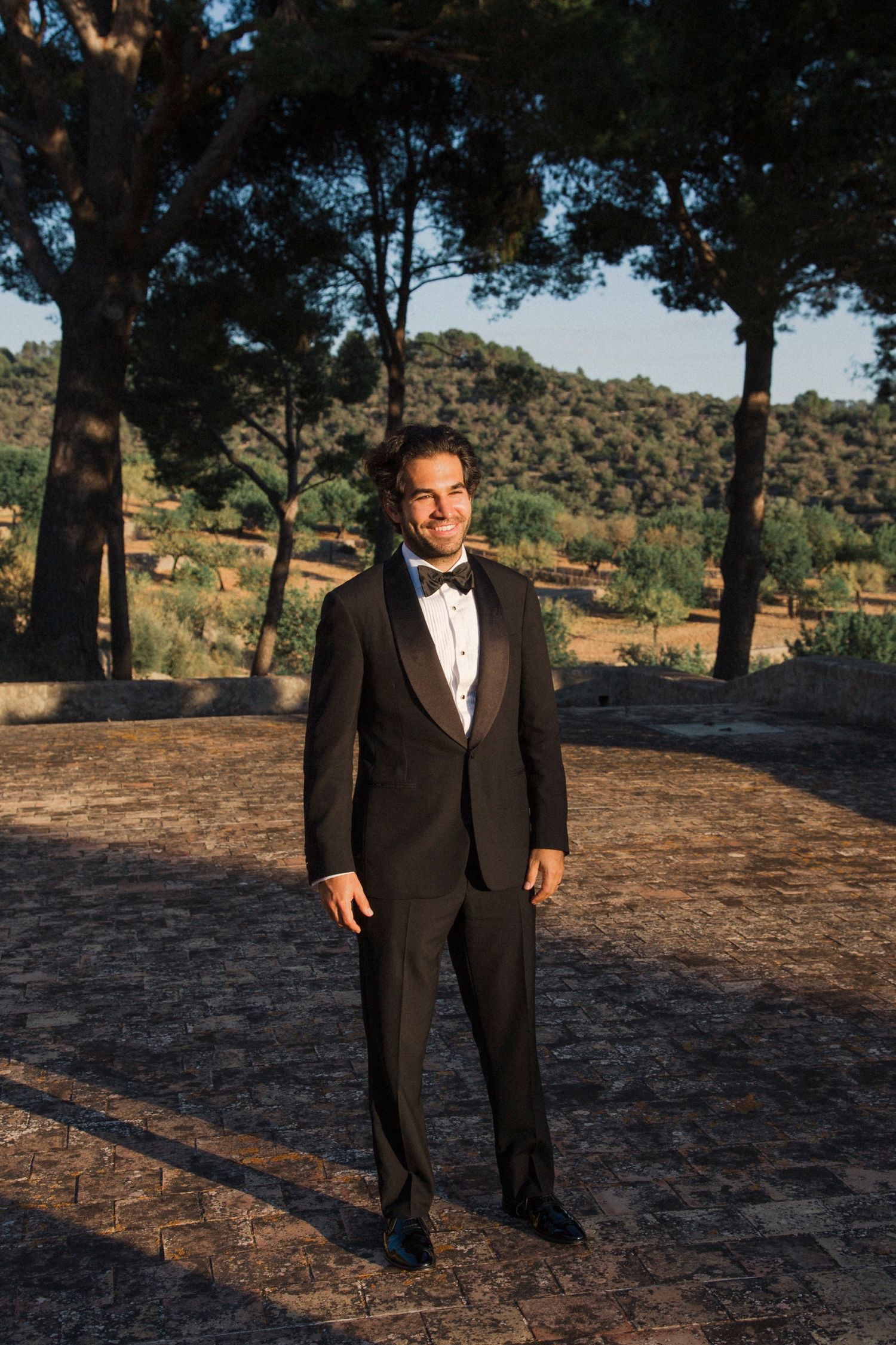 A person in a black tuxedo and bowtie stands outdoors against a natural landscape with trees and hills.