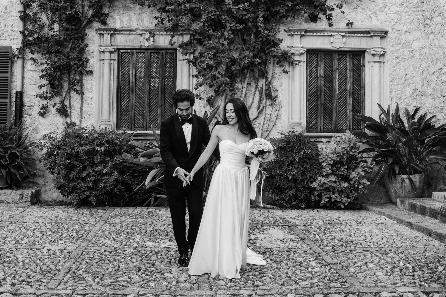 A black and white wedding photograph captured in front of a rustic building with climbing vines and cobblestone ground.