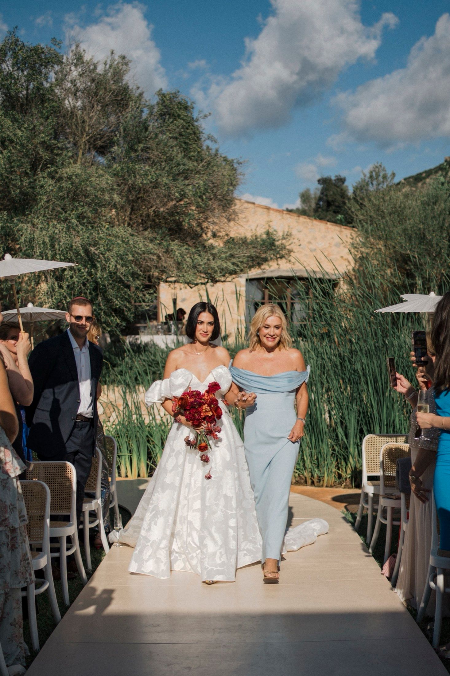 Wedding ceremony processional on a wooden boardwalk surrounded by tall reeds and blue skies on a sunny day.