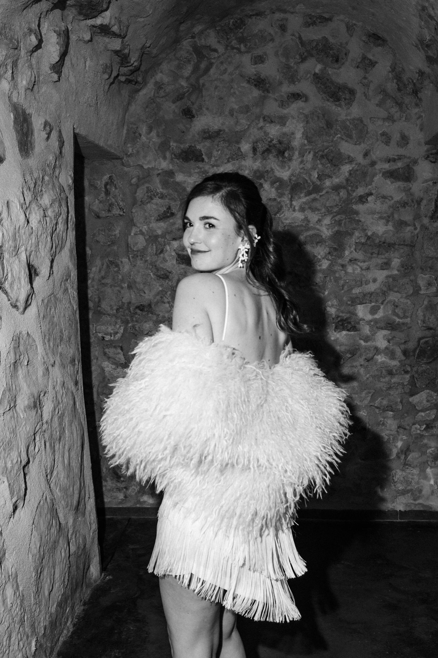 A glamorous black and white photo of someone in a feathered white party dress against a stone wall.