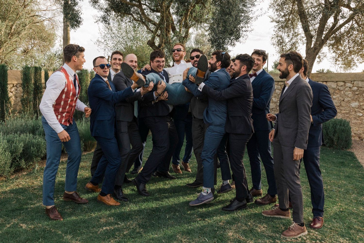 A large group of wedding guests gather outdoors under trees for informal photos.