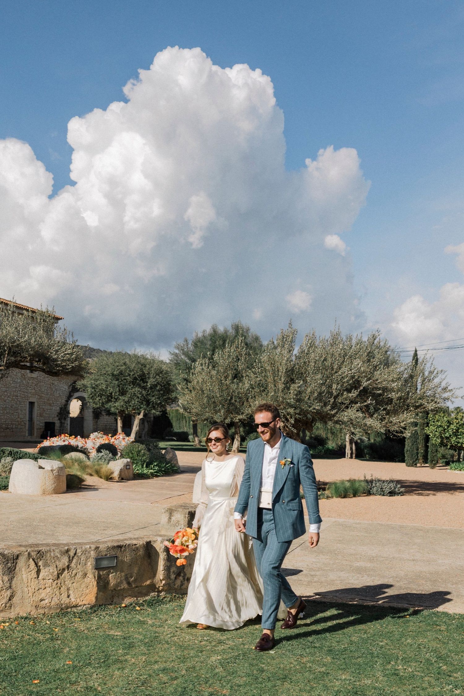A couple walks along a scenic Tuscan hillside path lined with olive trees on a sunny day with white puffy clouds.