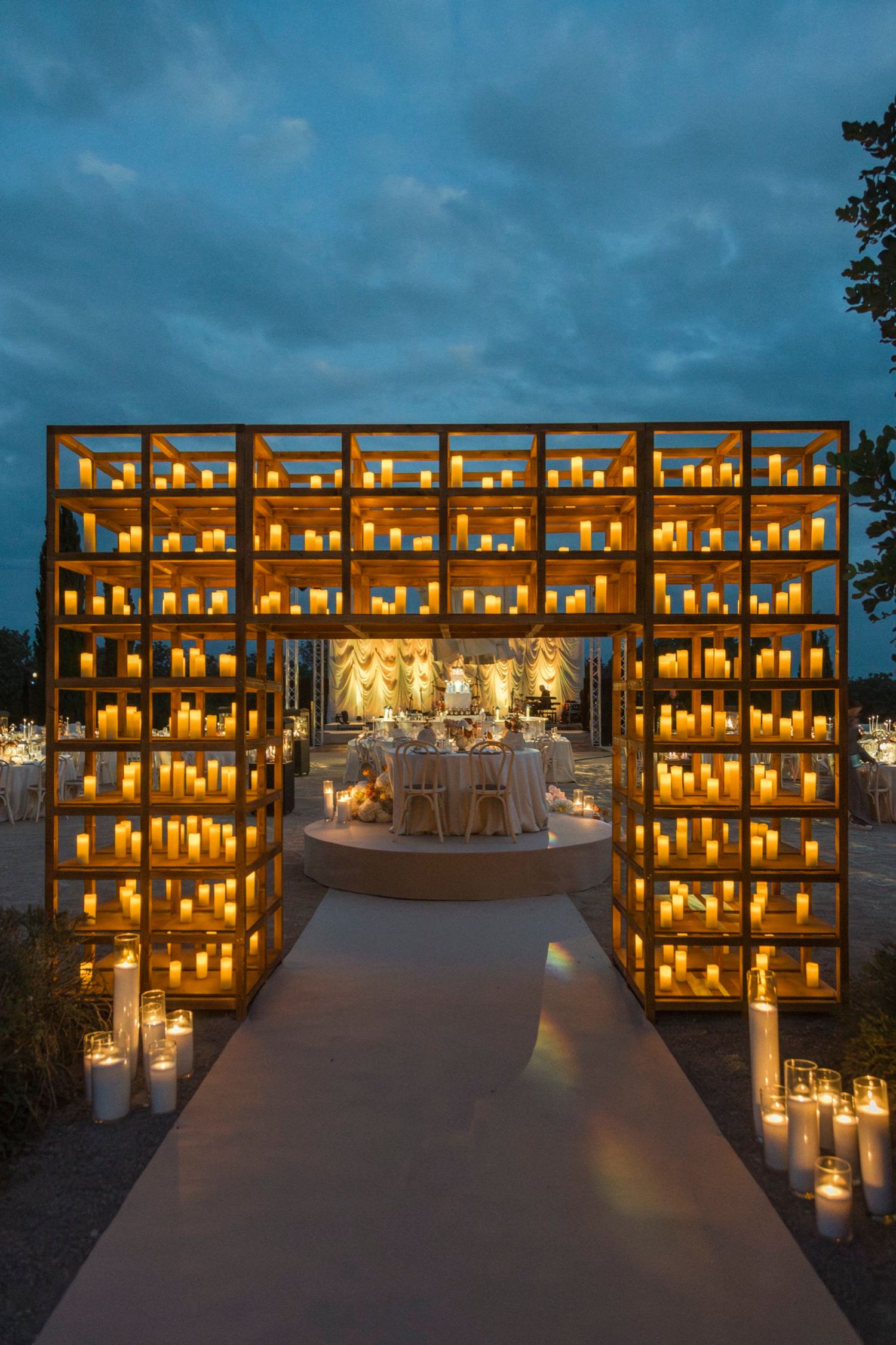 Illuminated wooden shelves with glowing candles create a serene memorial display against a dusky blue sky.