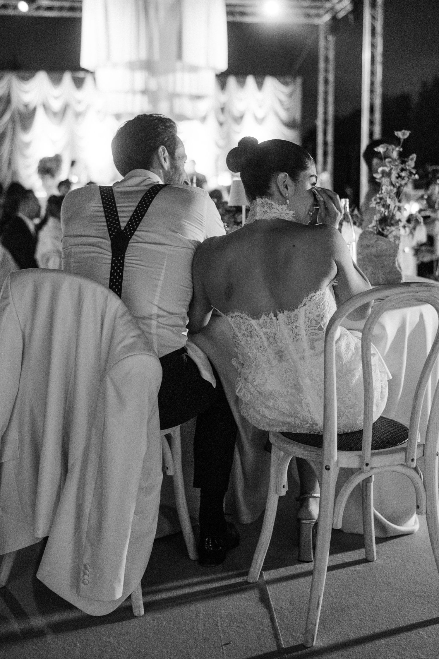 Wedding guests seated in white chairs at an elegant reception venue with decorative lighting and white tablecloths.