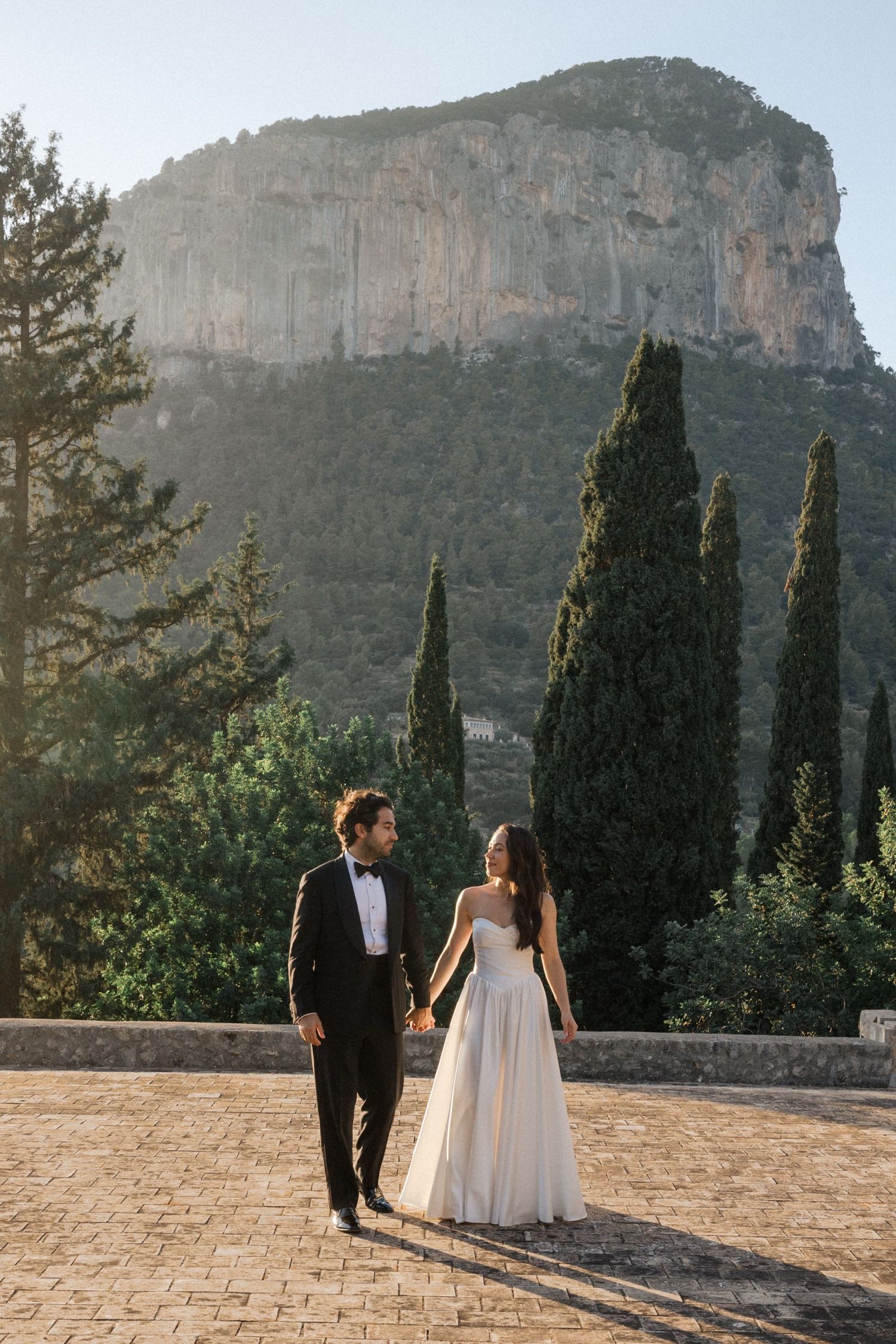 Couple walking along stone pathway with dramatic mountain cliffs and cypress trees in background at sunset.