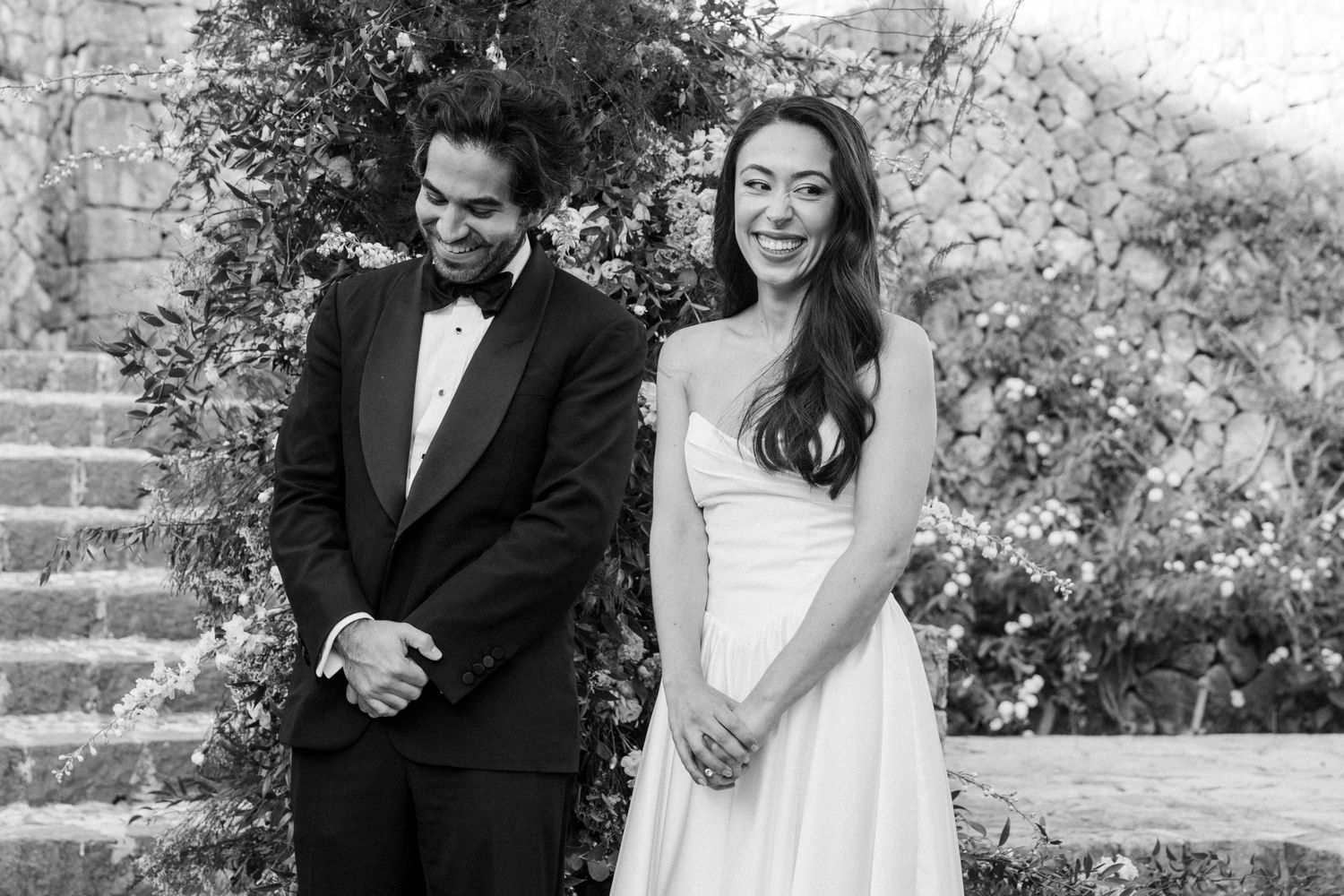 A happy couple laughs together during an outdoor wedding photo session in a garden setting.