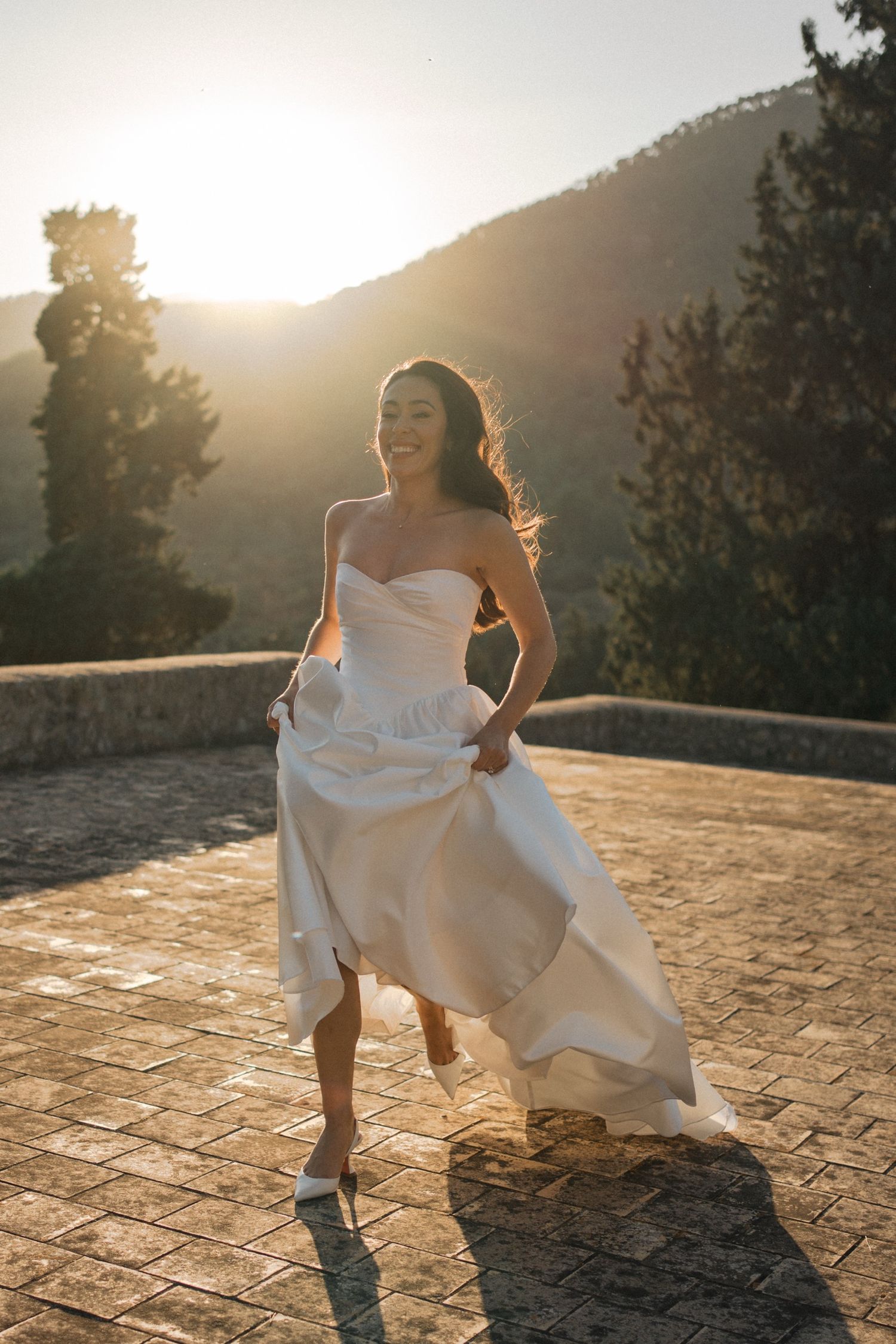 A person in a flowing white wedding dress and cowboy boots dances joyfully at sunset on a stone patio.