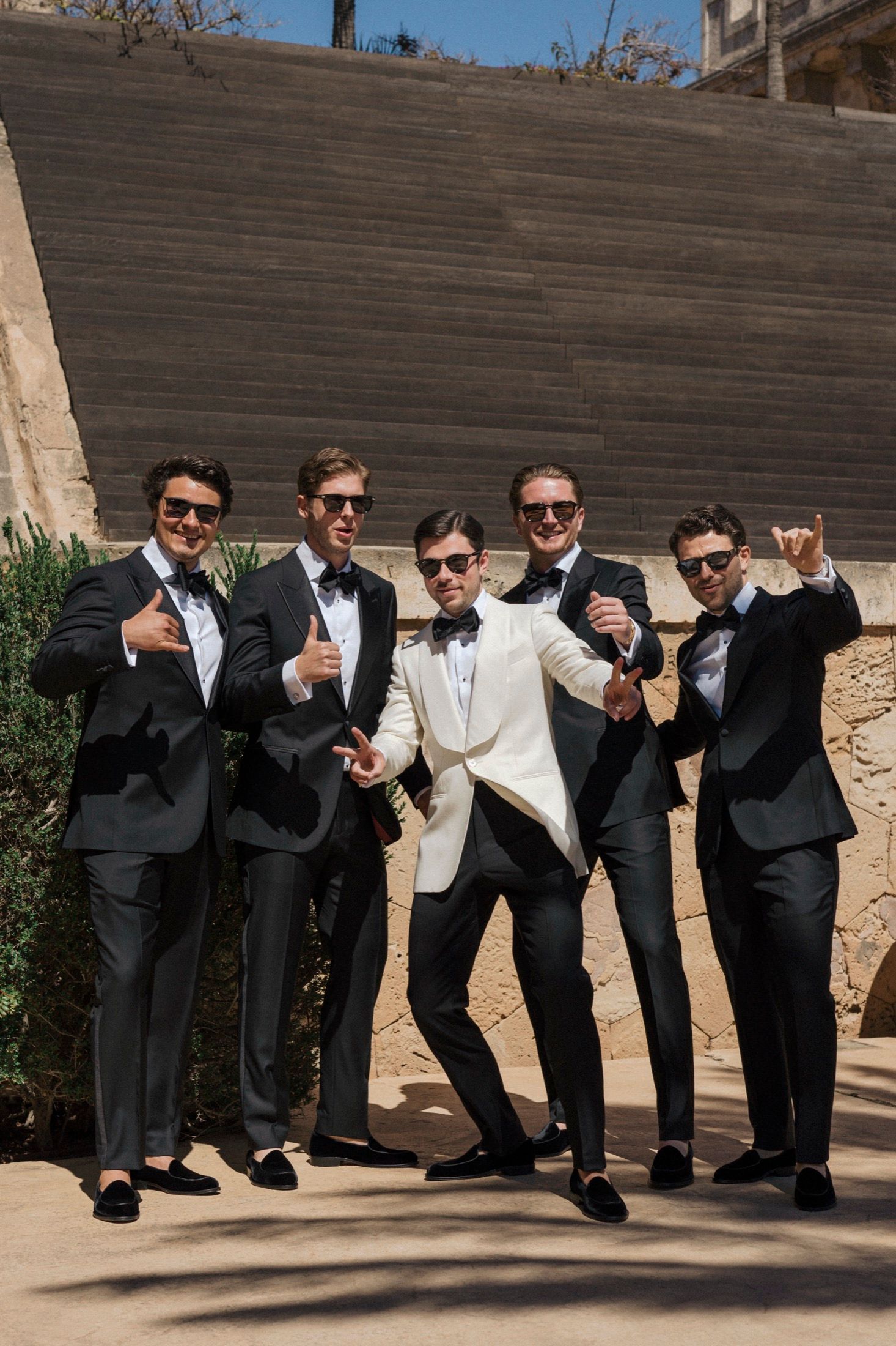 Five men in black tuxedos and sunglasses pose confidently outdoors, with one man in a white jacket centered.
