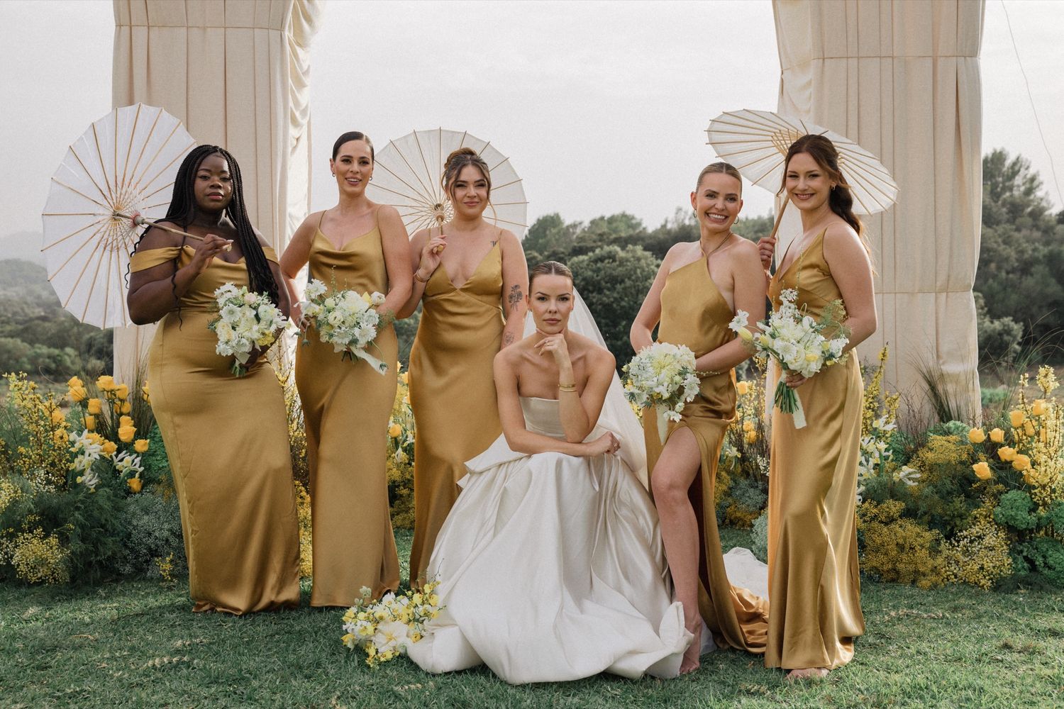 Wedding party poses under an elegant archway with bridesmaids in gold dresses holding white parasols.