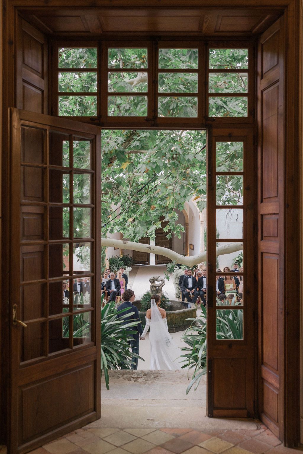 A wedding ceremony viewed through ornate wooden doors framing a garden setting with guests.