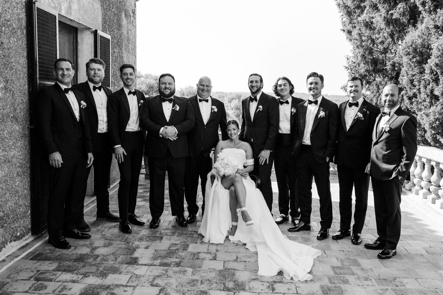 Wedding party in formal black attire poses together on a stone terrace.